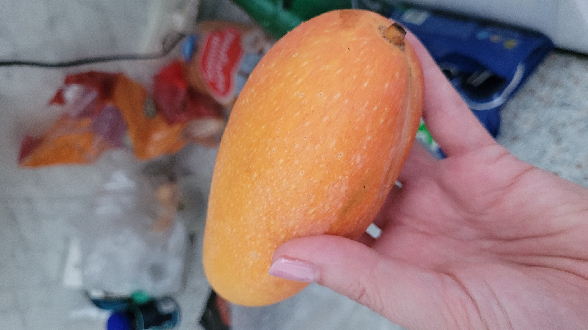 A mango is being held in a person's hand. The mango is orange in color and has a stem at the top. The person's hand has pink nail polish. The background includes a bag of food and a blue package. The location is an indoor setting.