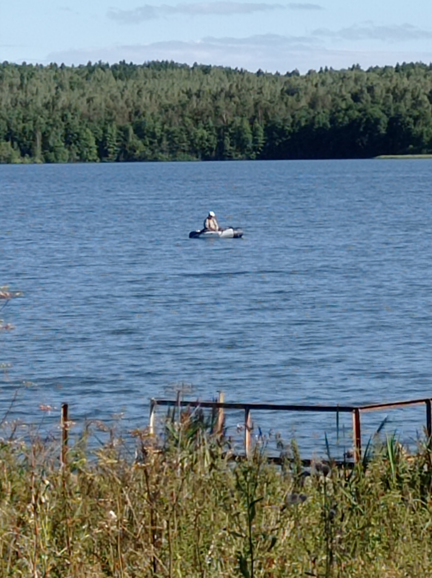 A person wearing a white hat and a light-colored jacket sits in a small, inflatable boat on a calm blue lake. The boat is positioned towards the left side of the frame. In the background, a dense, green forest lines the distant shore under a hazy blue sky with scattered clouds. In the foreground, tall, dry grasses and reeds are visible, obscuring a portion of a wooden and metal dock. The scene appears to be a recreational area in Degtyarsk, Russia, likely a park or natural reserve.