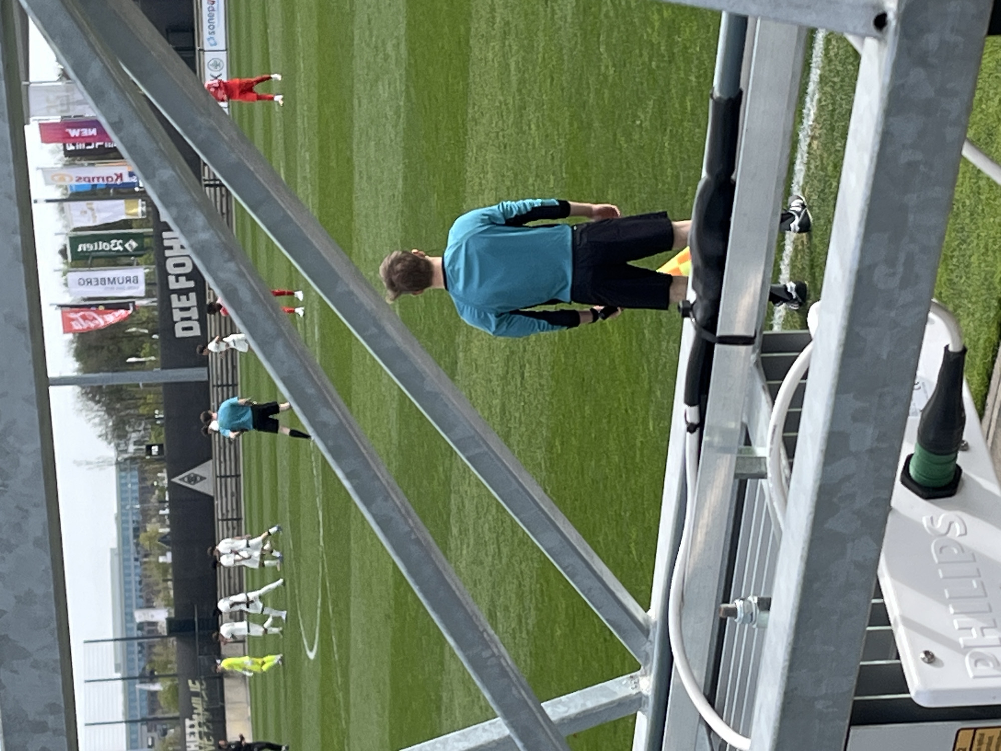 The image shows a partial view of a soccer field from an elevated position, likely a camera tower. In the middle ground, there are several soccer players on the field; most wear white jerseys and shorts with black socks, while one wears a bright yellow jersey and black shorts. Another player wears a red jersey and shorts. Further away on the field, a person in a blue long-sleeved shirt and black shorts is standing near the sideline, appearing to be an official or coach. The perspective is partially obscured by the metal structure of the tower. In the background, banners and signs are visible, including one that reads "DIE FOHLEN". The city of Mönchengladbach, Germany, is also apparent from the architecture and landscaping visible in the distance. A Philips lighting fixture is attached to the tower structure near the right side of the image.