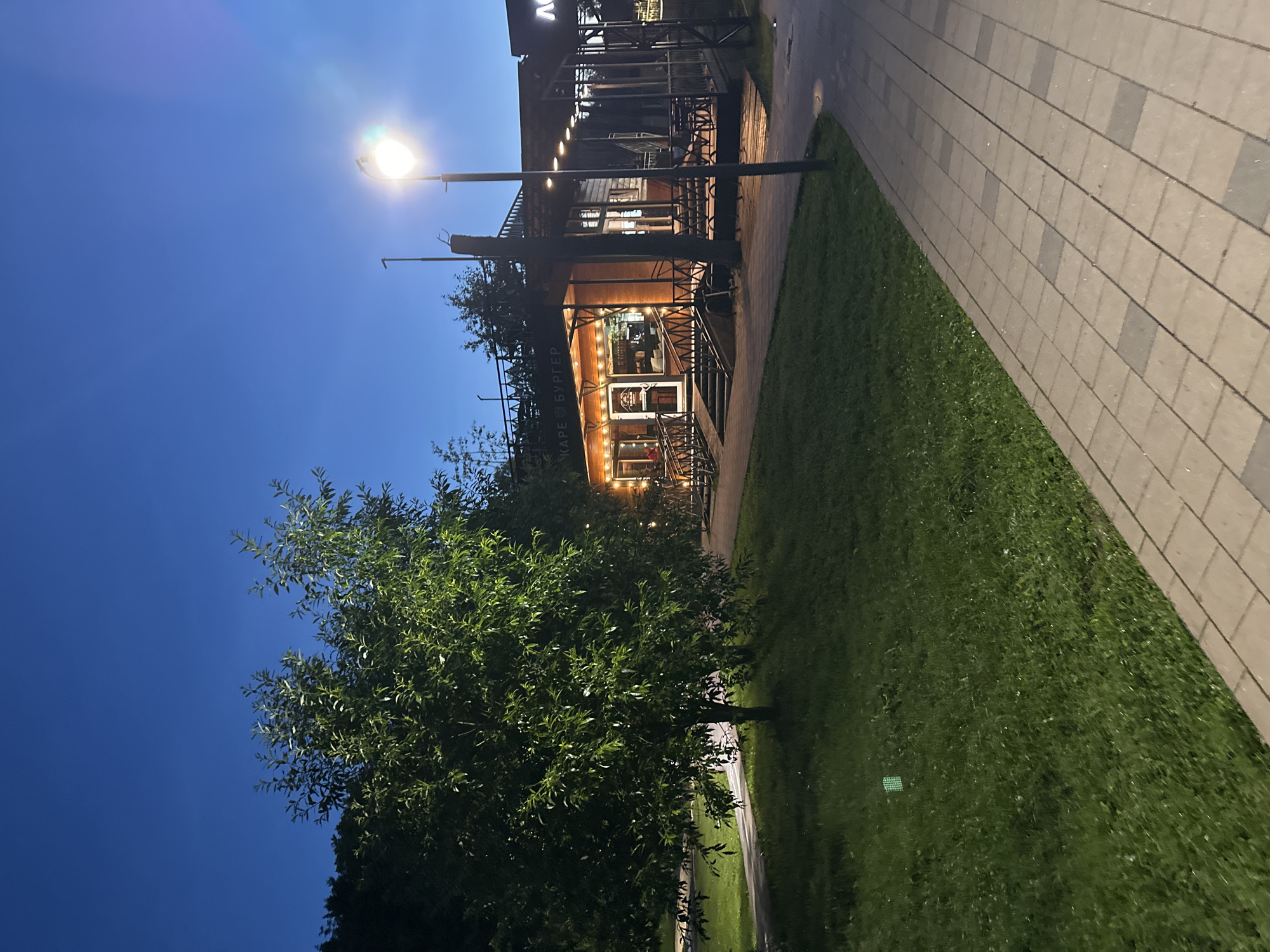 The image depicts a street scene at dusk in Solnechnogorsk, Russia, specifically near a cafe called "КАРЕ БУРГЕР" which translates to "Square Burger". A well-lit, wooden cafe with outdoor seating and a covered patio is the central focus. The cafe is surrounded by greenery, including a large tree in the foreground to the left and manicured lawns. A paved walkway runs along the right side of the image, leading away from the cafe. A streetlamp illuminates the scene, casting a warm glow on the path. The sky is a deep blue, indicating the transition from day to night. No people are visible in the image.