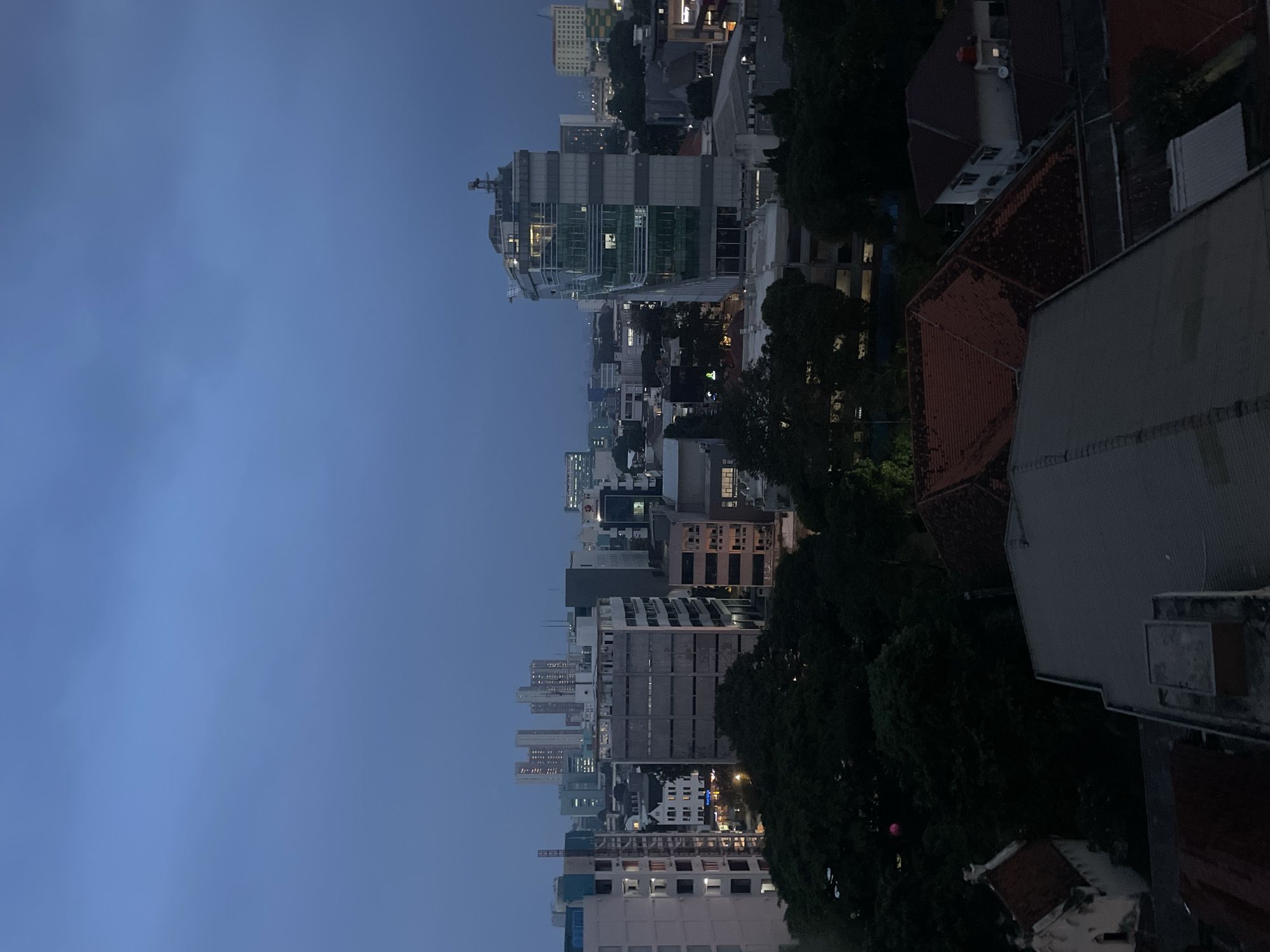 The image shows a cityscape of Central Jakarta, Indonesia, at dusk or twilight. The sky is a deep blue, with some clouds visible. The city is filled with buildings of various heights and architectural styles, including modern skyscrapers with glass facades and older, more traditional structures. The lights of the city are beginning to turn on, casting a warm glow against the darkening sky. In the foreground, there are several rooftops of residential or smaller commercial buildings, some with red tiled roofs. In the mid-ground and background, there are taller office buildings and apartment complexes. Trees are scattered throughout the city, adding a touch of greenery to the urban landscape. One building, in the mid-ground right, has a prominent glass facade with visible internal lighting. The overall impression is one of a busy and vibrant city as it transitions from day to night.