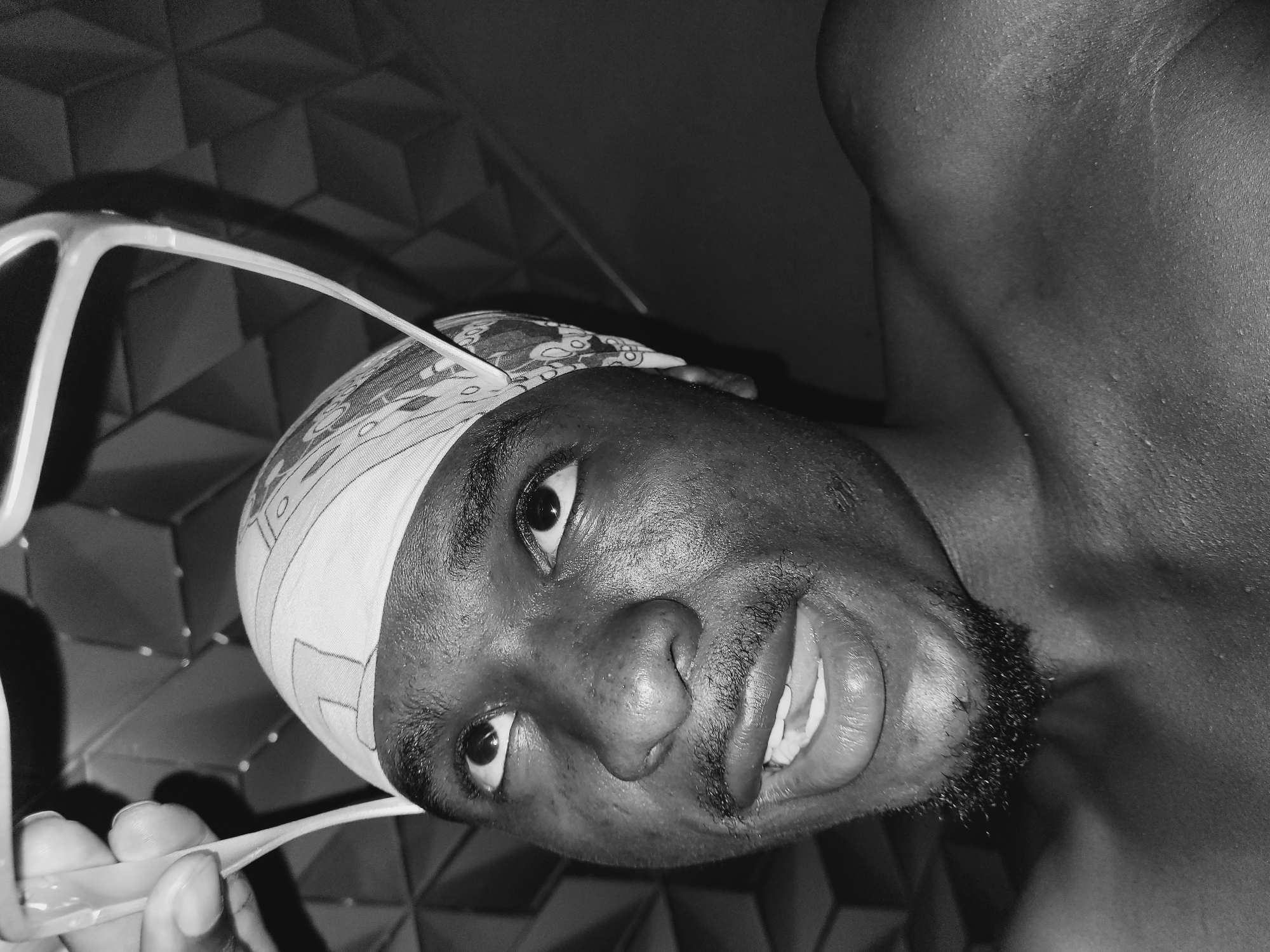 A close-up, low-angle, black and white shot captures a young Black male from the chest up, looking upwards with his mouth slightly open. He is wearing a patterned bandana on his head. A pair of light-colored glasses is held above his head by a hand, the fingers gripping the plastic frame. His eyes are wide, and he has a light mustache and a small goatee. The background features a textured, geometric pattern that appears to be a wall covering, dimly lit. The lighting highlights the sweat on his skin, suggesting a warm environment, potentially within a residential setting in Bukuru, Nigeria.