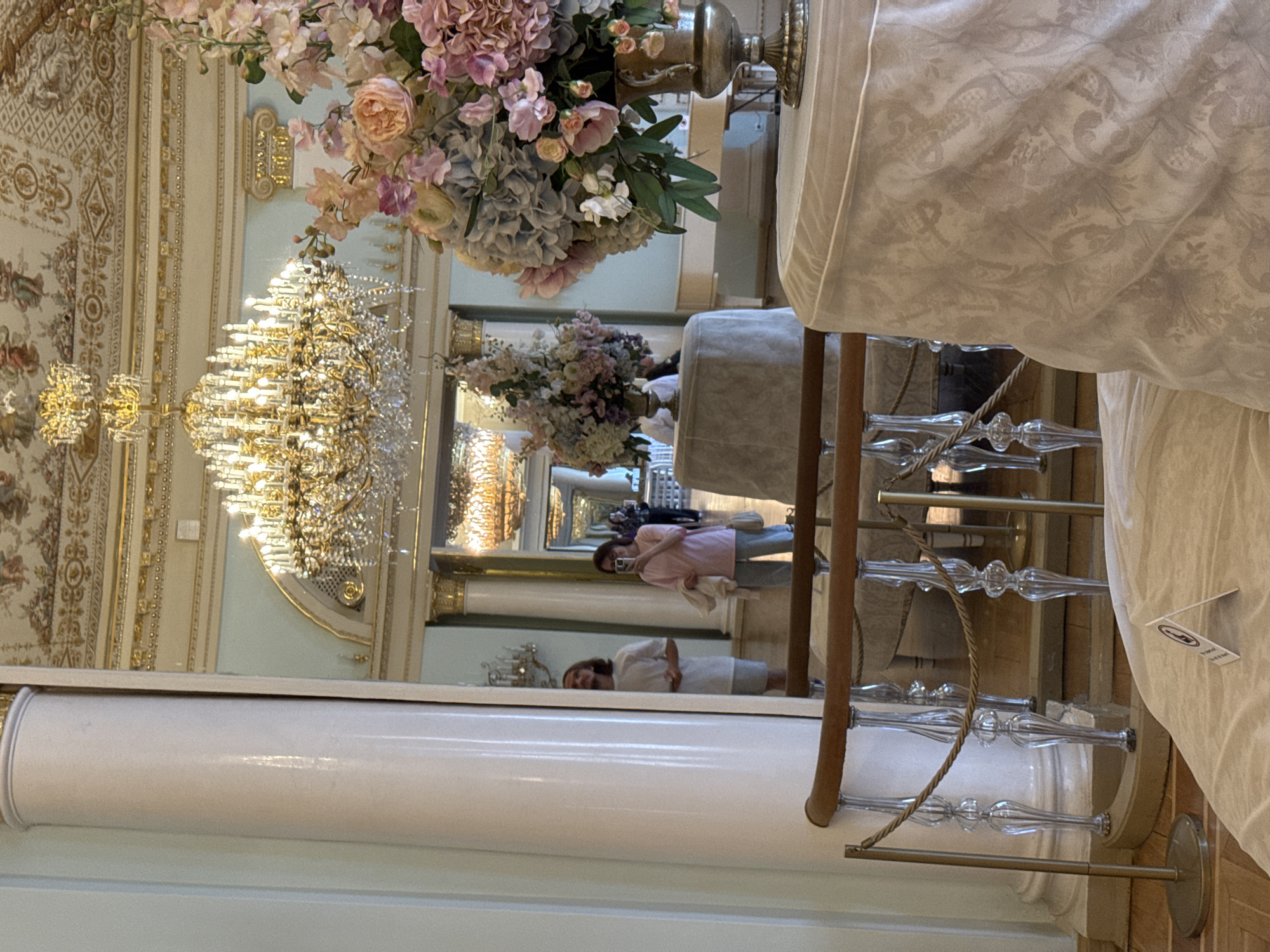 The image depicts the interior of a grand room in the Catherine Palace, located in Pushkin, a suburban town within the city of Saint Petersburg, Russia. The room is elaborately decorated with gilded architectural elements, including a highly ornate chandelier and intricate ceiling frescoes. In the foreground, a large floral arrangement of pink and blue flowers is displayed in a metallic vase on a draped table. A mirrored wall reflects the opposite side of the room, showing a large pillar and two individuals. One person, a woman in a pink top and grey pants, is taking a photo with her phone. Another person, a man in a white t-shirt and light shorts, is standing and looking upwards. The reflection also shows another chandelier in the distance, suggesting a series of interconnected opulent rooms. The overall scene conveys a sense of luxury and historical grandeur, characteristic of imperial palaces.