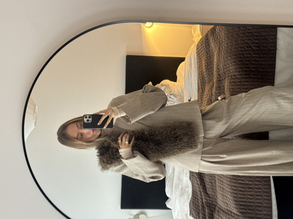 Location: Private residence, city of Unknown, Unknown.
Scene: A female subject is depicted in a full-length, arched mirror, taking a selfie in what appears to be a bedroom. The subject has light brown hair and is wearing a beige pinstriped pantsuit consisting of an oversized blazer and wide-leg trousers. A light beige turtleneck is visible beneath the blazer. A dark brown, fluffy faux fur stole is draped across her left shoulder and chest. She holds a black smartphone with her right hand, obscuring part of her face, and a ring is visible on her left ring finger. The background features a bed with a dark brown upholstered headboard, white sheets, and a brown quilted bedspread. A wall-mounted light fixture emitting a warm yellow glow is visible on the right, and a white, inverted bowl-shaped ceiling light is reflected at the top. The surrounding walls are light-colored.