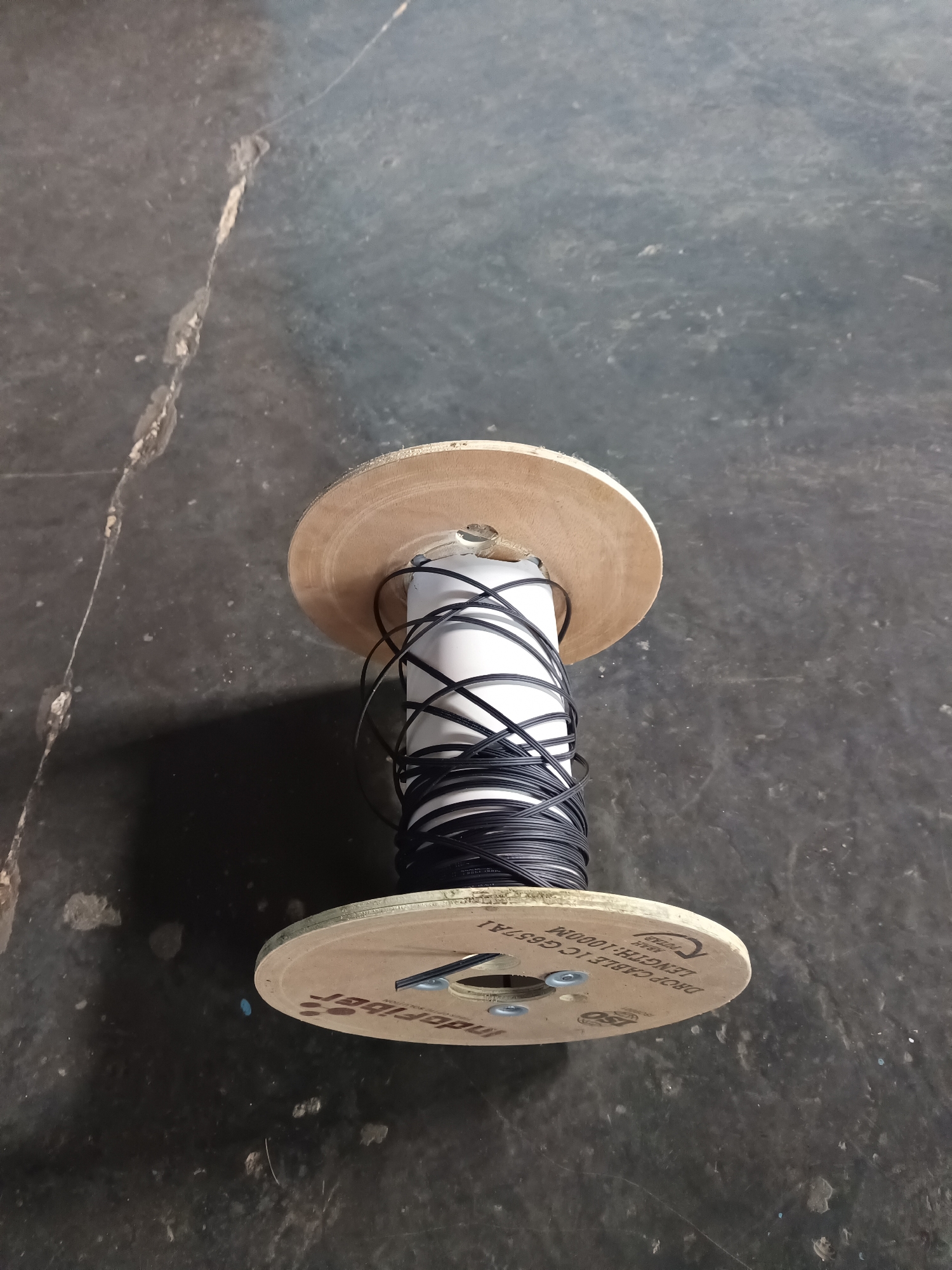 The image shows a spool of black wire, likely fiber optic cable, resting on a dark, textured surface that appears to be asphalt or concrete. The spool has wooden flanges with some text printed on them, including "DROP CABLE 1000M" and "ISO". The wire is wound tightly around a white cylindrical core. The scene is outdoors, as indicated by the natural lighting and the uneven texture of the ground. There are no people or animals visible in the image. The overall impression is one of raw materials or construction supplies. The location is identified as Terbanggi Marga, Indonesia.