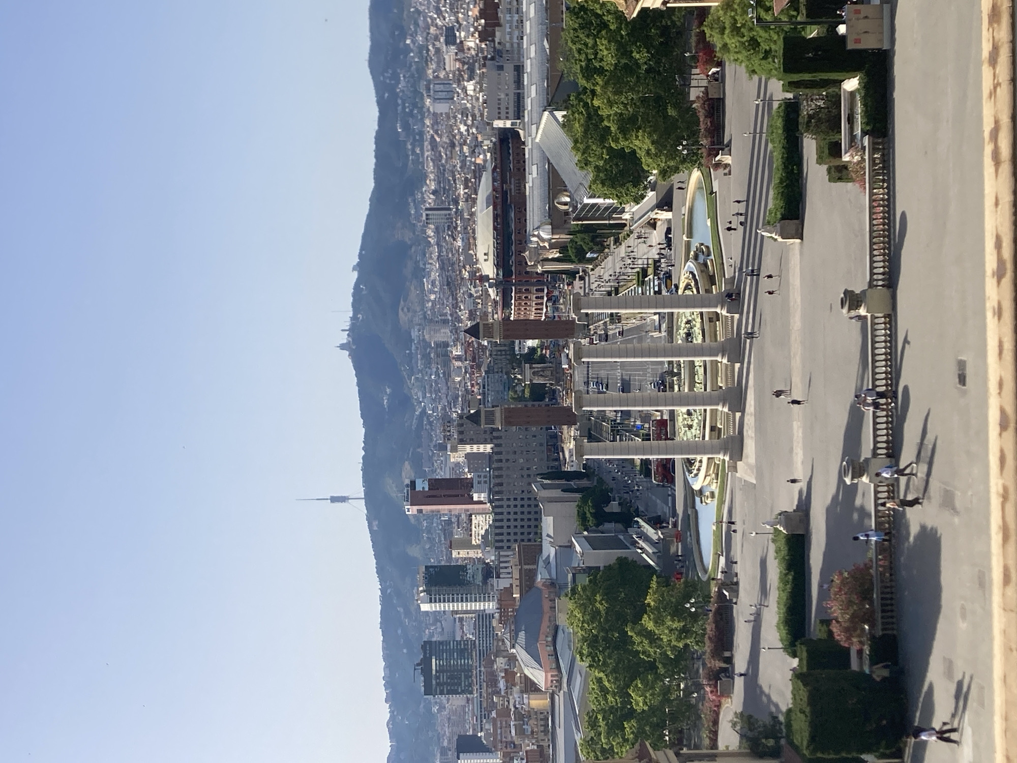 This is an eye-level, wide-angle shot of a panoramic view of Barcelona, Spain, looking towards the city skyline and a distant mountainous backdrop. In the foreground, there is a wide paved plaza with a balustrade. Several people are visible walking and standing on the plaza. Directly in front of the viewer are two large classical columns. Beyond these columns is a long fountain with water jets, and a wide avenue stretches into the distance.
The avenue is flanked by trees and buildings on either side. Prominent structures include the Palau Nacional on the left, with its distinctive domed roof, and on the right, the Arenas de Barcelona, a former bullring with a distinctive circular structure. Further into the city, various modern and older buildings are visible, including several high-rise towers. In the far distance, a prominent transmission tower, likely the Collserola Tower, stands on a hilltop, with another smaller structure on a separate peak. The sky is clear and blue. The lighting suggests it is daytime, likely morning or afternoon, given the distinct shadows.
