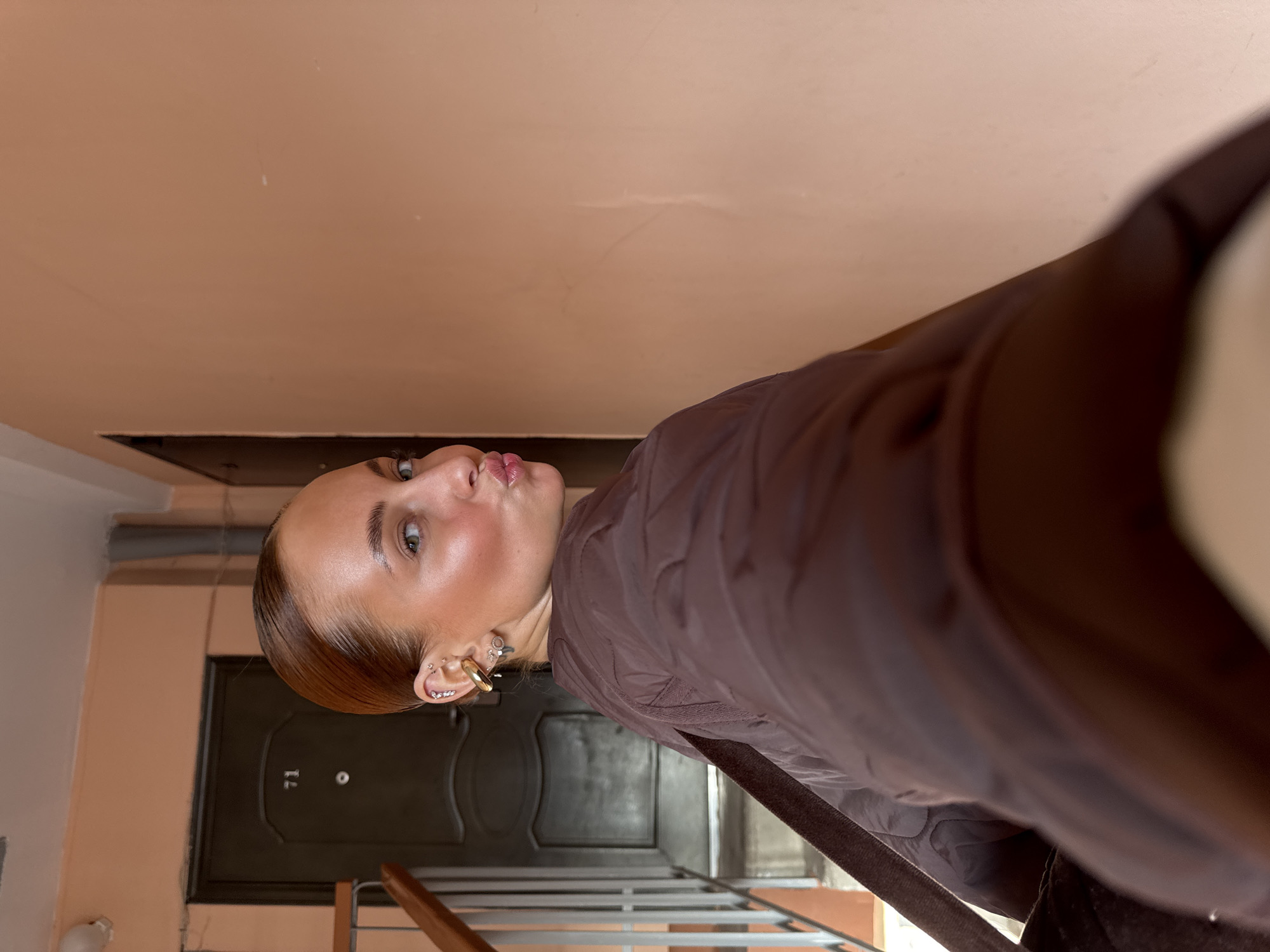 A woman with fair skin and light brown hair pulled back is pouting her lips towards the camera. She is wearing multiple earrings in her left ear, including a large gold hoop. She is wearing a brown quilted jacket.
The background suggests an indoor location, likely a stairwell or hallway in an apartment building. The walls are painted a light peach color. A dark-colored door with the number "71" is visible behind her, suggesting an apartment unit. A metal railing is visible on the right side of the frame.
The scene appears to be in Chaikovsky, Russia.