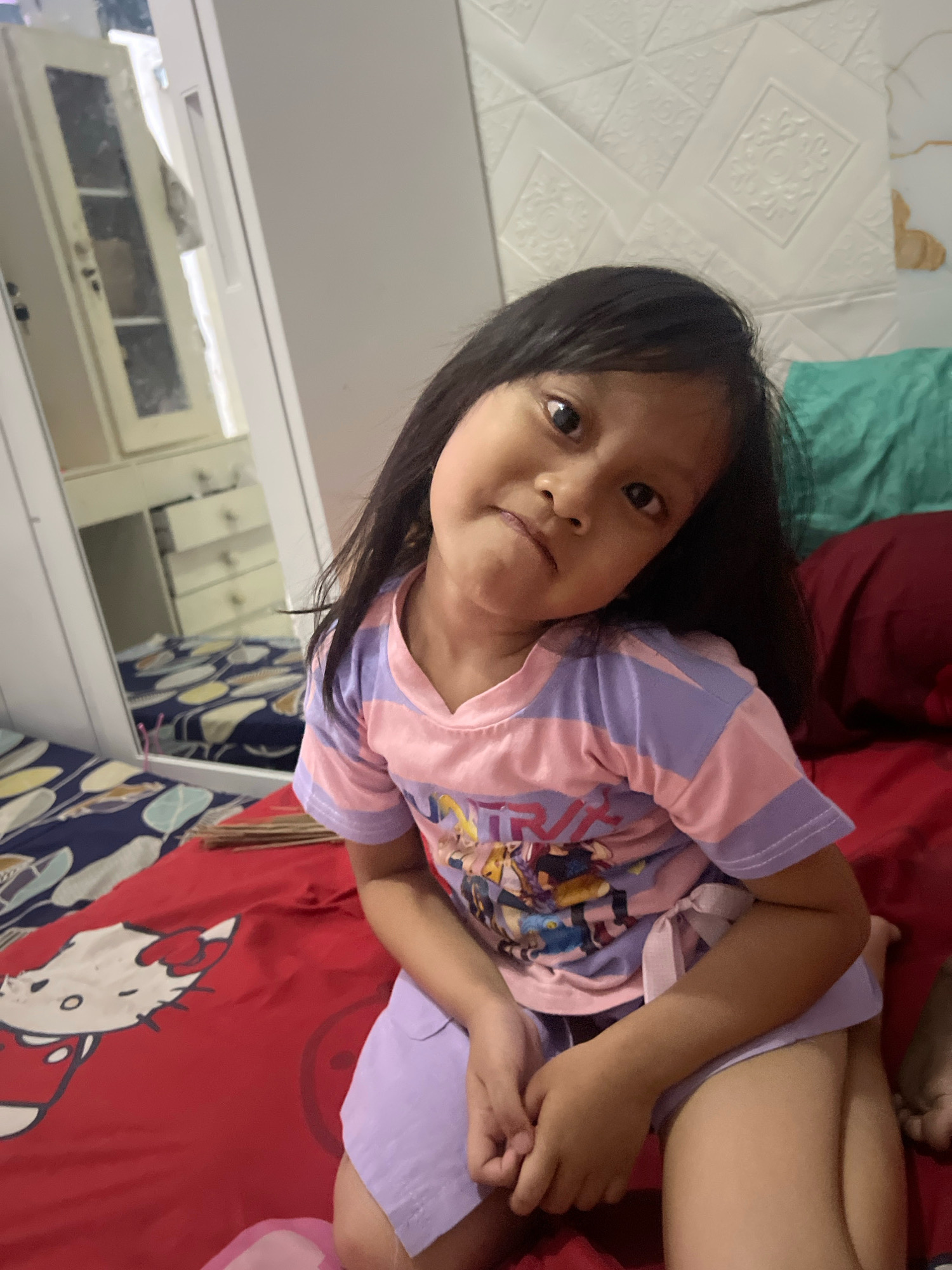 A young girl with dark hair and brown eyes is sitting on a bed. She is wearing a pink and purple striped t-shirt with an image of cartoon characters on it, and light purple shorts. Her head is tilted to her left, and she is looking at the camera with a slightly puckered expression. The bed has a red sheet with a Hello Kitty pattern and other patterned sheets visible. In the background, there is a white textured wall, a large white wardrobe with mirrors reflecting the room, and pillows in shades of teal and burgundy. The lighting suggests it might be daytime or evening indoors. The scene appears to be a bedroom in a home.