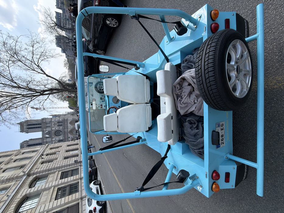 A light blue, open-top, two-door recreational vehicle, resembling an electric Moke, is observed from a slightly elevated rear perspective on a paved city street in Paris, France. The vehicle features white bucket seats in the front and a white bench seat in the rear. Black seatbelts are attached to the vehicle's prominent light blue tubular roll bar frame. The rear cargo area contains two rolled-up blankets, one gray and one light brown. A spare tire with a silver multi-spoke rim is mounted externally on the lower rear. A French license plate, "268-EG," is affixed to the rear panel, beneath which "THEMOKEENRIDE.COM" is partially legible.
In the mid-ground, directly ahead of the vehicle's implied direction of travel, the iconic Gothic towers and rose window facade of Notre Dame de Paris are clearly visible, with a construction crane positioned to its right. To the left of the street, a multi-story classical Parisian building with numerous windows is present, alongside several parked cars, including a white hatchback. To the right, a large deciduous tree with bare branches stands, followed by more urban buildings and a lane of traffic, which includes a black sedan. Scattered pedestrians are visible on sidewalks in the background. The scene is illuminated by bright daylight under a clear blue sky.