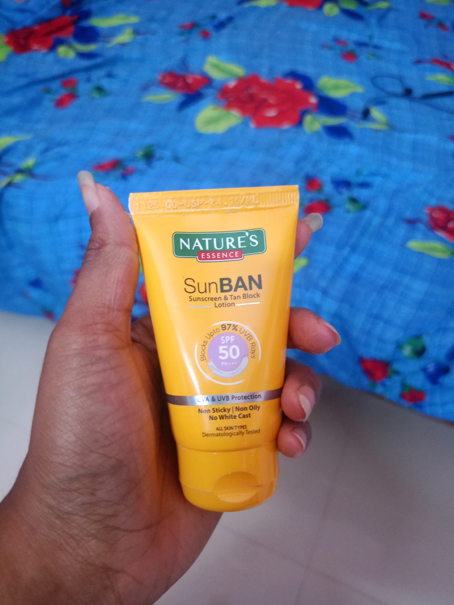A person's hand holds a yellow tube of "NATURE'S ESSENCE SunBAN Sunscreen & Tan Block Lotion" in front of a blue fabric with a floral print. The tube is oriented vertically, with the cap at the top. The label on the tube is visible, showing the brand name, product name, SPF 50 rating, and other details. The hand is positioned to obscure a portion of the tube's label. The background is out of focus, showing a blue fabric with a repeating pattern of red and green flowers. The lighting appears to be natural. The image is taken from a slightly high angle, looking down at the hand and the product.