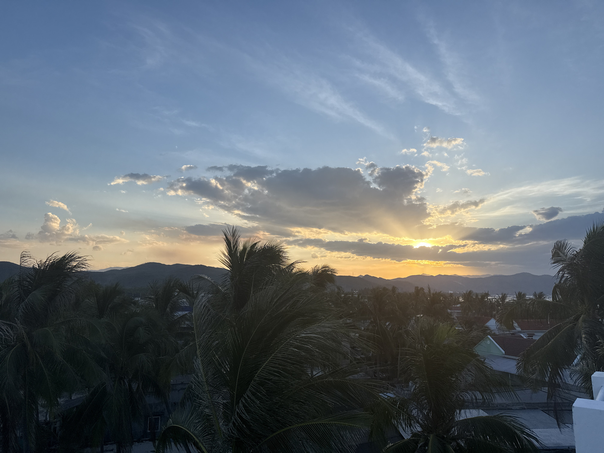 A high-angle, wide shot captures a tropical sunset over a coastal town. The sky transitions from a pale blue at the top to a vibrant orange-yellow near the horizon. Wispy, cirrus clouds are scattered across the upper sky, while thicker, more dramatic clouds are clustered near the setting sun, casting rays of light downwards. Below the sky, a dense canopy of palm trees dominates the foreground, their fronds silhouetted against the warm light. Peeking through the trees are the rooftops of houses, some with tiled roofs in shades of red and green, indicative of a warm climate. In the distance, layered mountains form a dark silhouette against the glowing horizon, with the faint outline of the ocean visible to the right. The overall impression is one of serene, natural beauty and a peaceful evening in a tropical setting. There are no people or animals visible in the scene.