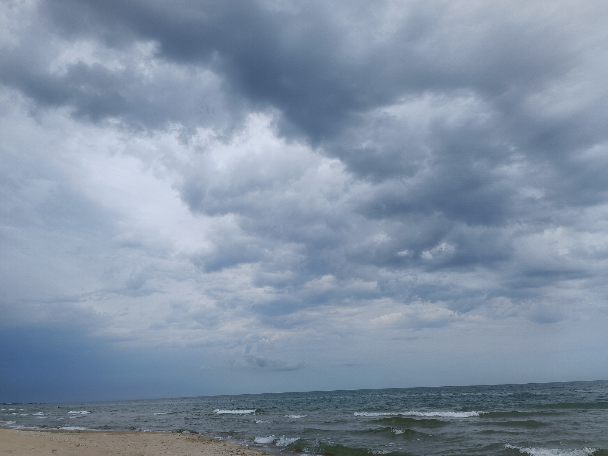 The image depicts a beach scene with a stormy sky. The foreground shows a sandy beach leading to the ocean. The water is a dark, choppy gray, with small waves breaking near the shore. The horizon is visible, separating the sea from the sky. The sky is filled with heavy, dark gray clouds, indicating a storm. There are no people or objects clearly visible on the beach or in the water, though a faint line of structures can be seen in the distance along the coast. The overall atmosphere is somber and overcast.