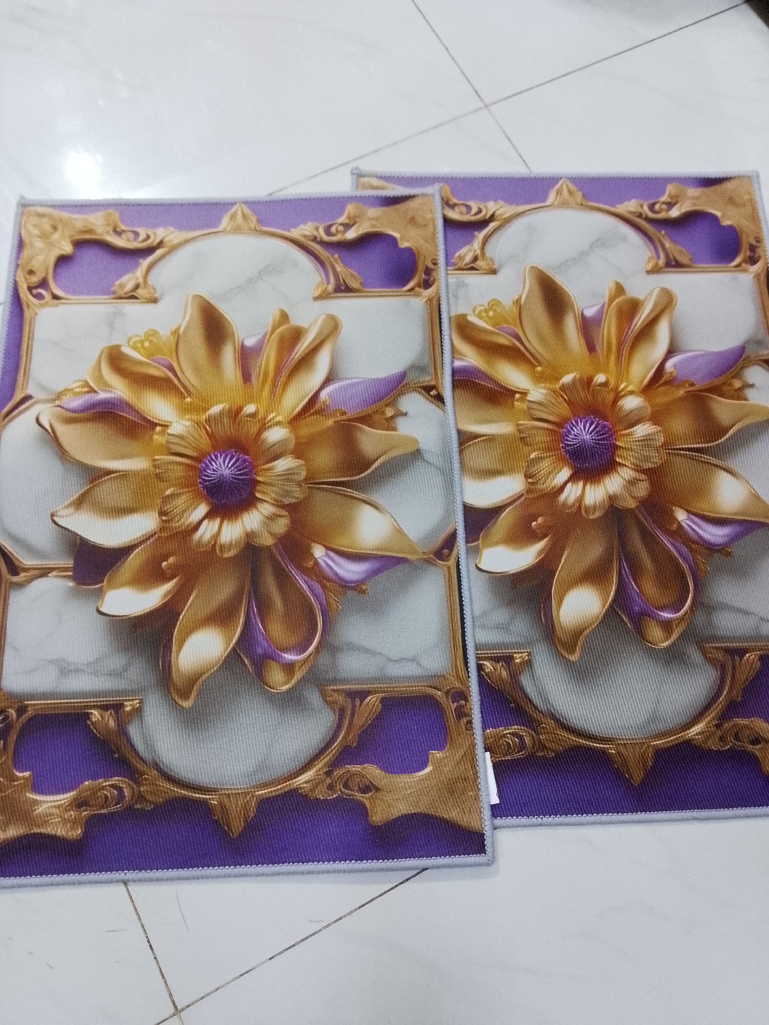The image presents two identical rectangular mats laid out on a light-colored, tiled floor, characteristic of an indoor setting. The mats are positioned side-by-side, with a slight overlap, suggesting they might be a pair.
Each mat features a vibrant and ornate design. At the center of each mat is a prominent, three-dimensional-effect flower, rendered in lustrous golden hues with striking purple accents on its petals and a densely textured purple core. The petals exhibit varying shades of gold, creating depth and a metallic appearance. This central floral motif is encircled by an elaborate, ornate golden frame, reminiscent of baroque or rococo artistry, set against a background that mimics white or light gray marble with subtle veining. The entire decorative area is bordered by a solid band of deep purple, which in turn has a subtle, light-colored stitched edge.
The floor beneath the mats is composed of light-toned, possibly white or off-white, square tiles with visible dark gray grout lines forming a grid pattern. The lighting in the scene is bright and even, illuminating the mats without casting harsh shadows, typical of indoor ambient or artificial light. There are no people visible, and no specific activity or event is taking place; the image is a static display of the mats. The mats appear new and clean. There is no visible text or branding on any part of the image.
The location context provided is Davao City, Philippines, although no specific geographical cues are discernible within the image itself.