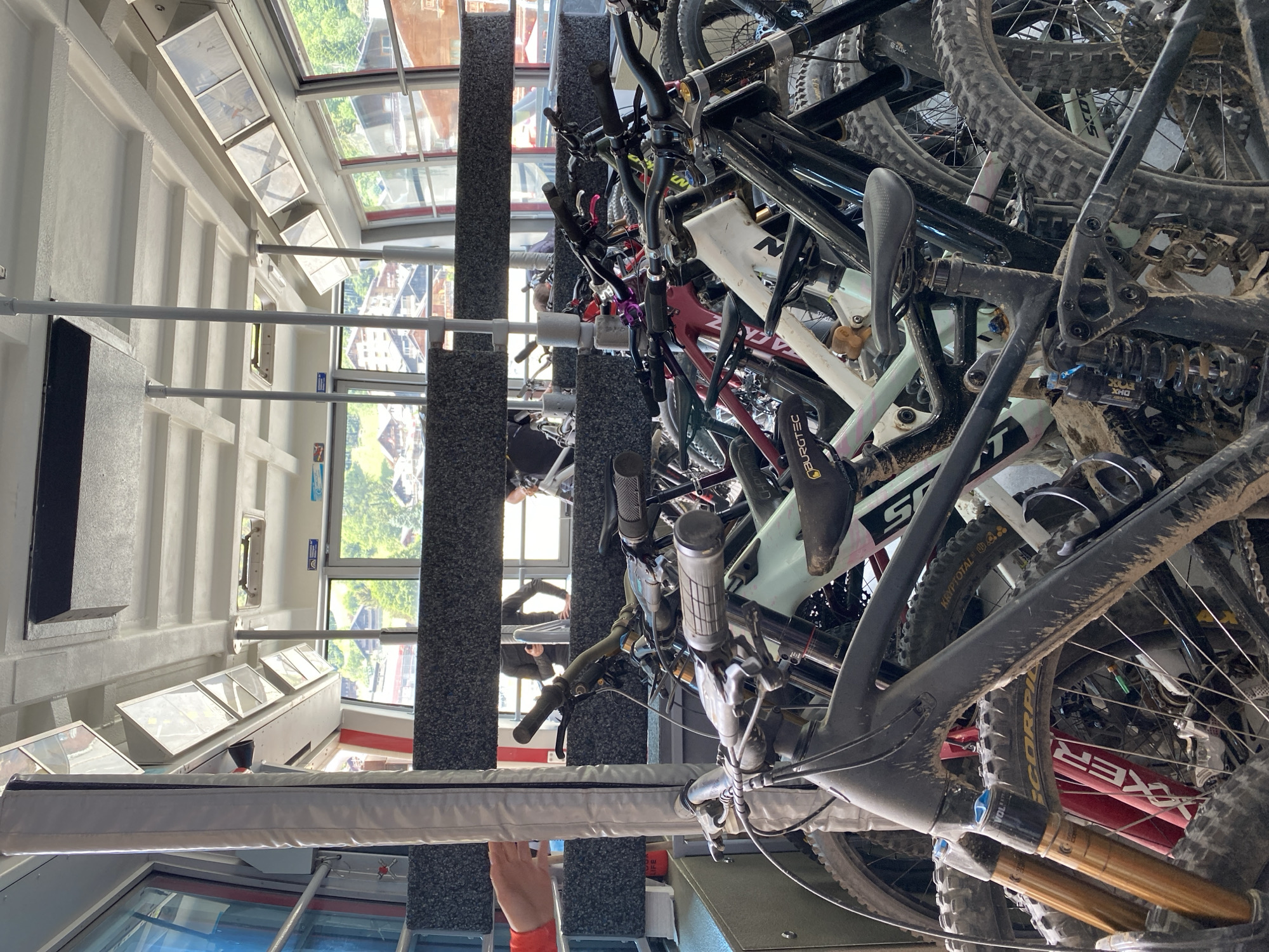 The image depicts a rack overflowing with mountain bikes inside a building, likely a cable car station, as suggested by the interior view of Champéry, Switzerland, visible through the windows. The bikes are piled haphazardly, showing signs of heavy use with mud and dirt covering their frames, wheels, and components. The color palette is predominantly dark, dominated by the black and gray of the bicycles. The environment is a mixture of industrial and outdoor elements, with the interior of the cable car station juxtaposed against the green landscape outside. Evidence of human activity is apparent, with people reflected in the windows in the background, although their actions cannot be explicitly determined.
