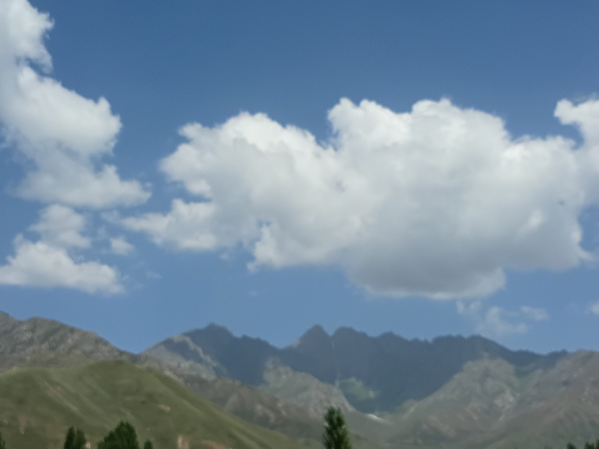 The image depicts a wide landscape under a bright blue sky with scattered white clouds. In the foreground, lush green rolling hills lead up to a range of jagged, rocky mountains in the background. Several trees are visible at the base of the hills. The scene is tranquil and suggests a rural or mountainous environment. The provided text states that the location is the city of Kalaynay, Tajikistan, which would place this scene within that geographical context. There are no people or specific actions depicted in the image; it is a static landscape view.