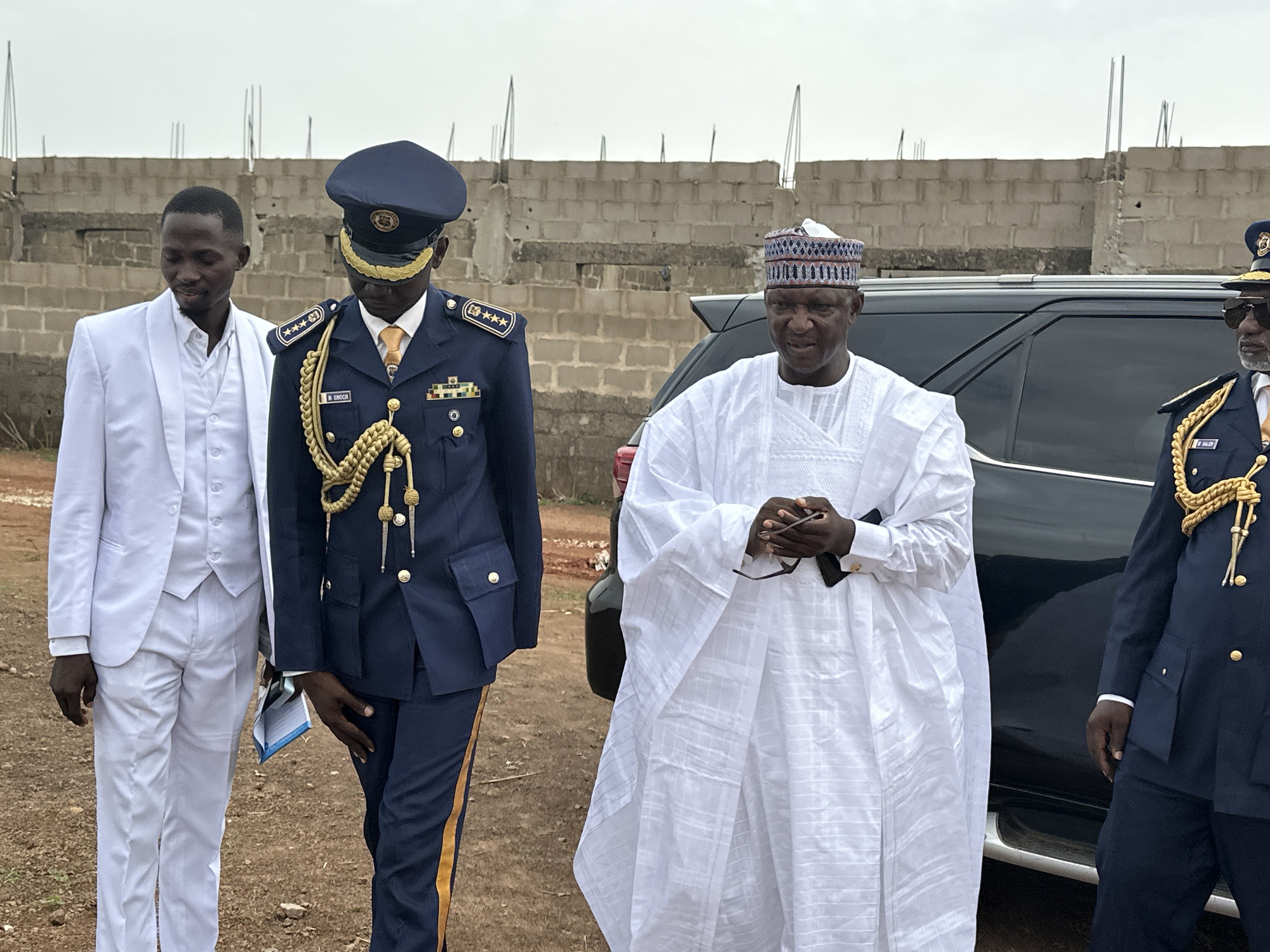 In Goni Gara, Nigeria, four men are standing outdoors on an overcast day. The man on the left wears a white suit. Next to him is a man in a dark blue military-style uniform with gold trim and a peaked cap. Another man wears a white traditional robe and a patterned cap. The fourth man, partially visible on the right, is also in a similar uniform to the second man. A black vehicle is behind the man in white robes. An unfinished concrete block structure is visible in the background.