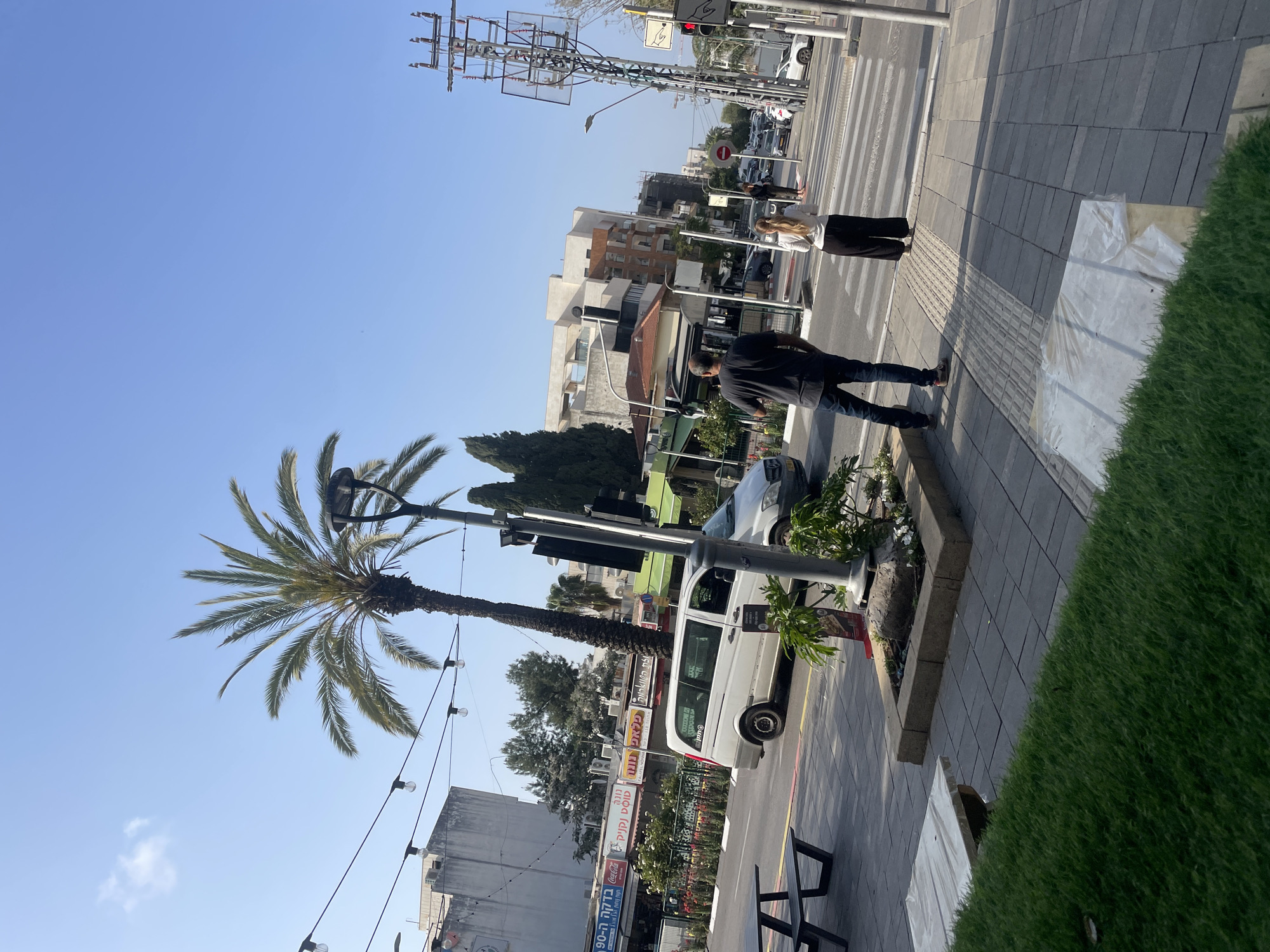 The image captures a street scene in Raanana, Israel. In the foreground, a palm tree stands adjacent to a sidewalk. A man in dark clothing is on the sidewalk, standing close to a planter. Further away, a woman is walking across a crosswalk. Cars are parked along the street, including a white van. Buildings and signage are visible in the background, along with utility poles and streetlights. The sky is blue.