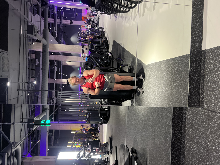 A muscular man in a red tank top and gray shorts flexes his arm while holding a phone, presumably taking a selfie. He stands in front of a large gym mirror that reflects a full view of the gym. Various exercise machines are visible in the background, and the gym has a modern design with overhead lighting and a speckled floor.