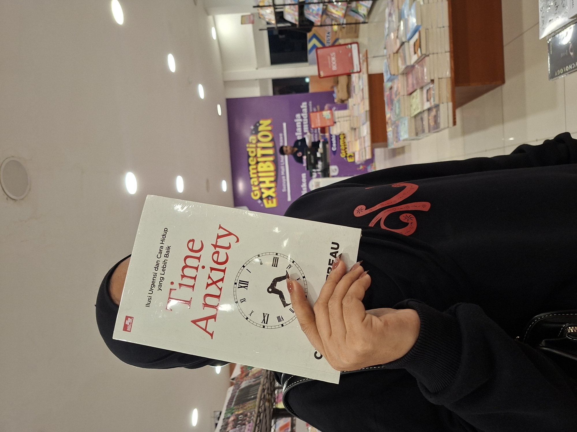 A woman wearing a black hijab and a black long-sleeved shirt with a red pattern is holding a book titled "Time Anxiety: Ilusi Urgensi dan Cara Hidup yang Lebih Baik" in front of her face. The book cover features a clock. The woman's right hand is visible. The photo appears to be taken indoors at a bookstore, with a "Gramedia EXHIBITION" sign visible in the background. A "BOOKS" sign, bookshelves filled with books, and a cashier area are also in the background. The sign indicates the location is at Suzuya Mall Lhokseumawe.