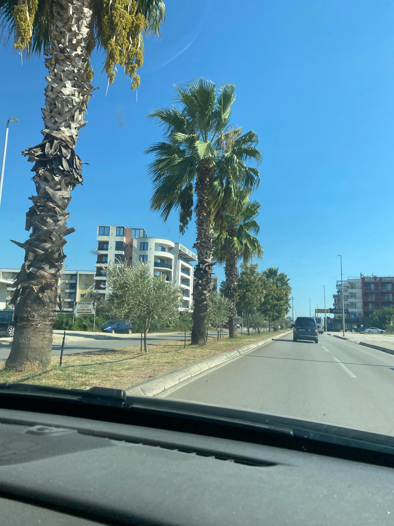 The image is a daytime, exterior, eye-level shot from inside a vehicle, looking forward through the windshield. The scene is a street in the city of Ulcinj - Ulqin, Montenegro, characterized by a clear blue sky and bright sunlight. The road is paved and appears to be a multi-lane highway with a visible lane divider.
Prominently featured on the left side of the road are several large palm trees with their characteristic rough trunks. Beyond the palm trees, there are modern multi-story buildings with balconies and windows. One building has a curved facade. An olive tree is also visible in this area, along with a street light. A dark-colored minivan is driving away from the camera in the right lane of the road. Further down the road, more buildings are visible, including one with a sign that reads "HOTEL EVROPA." The environment suggests a coastal or Mediterranean setting. The foreground of the image shows the dashboard of the vehicle, including the windshield wiper assembly.