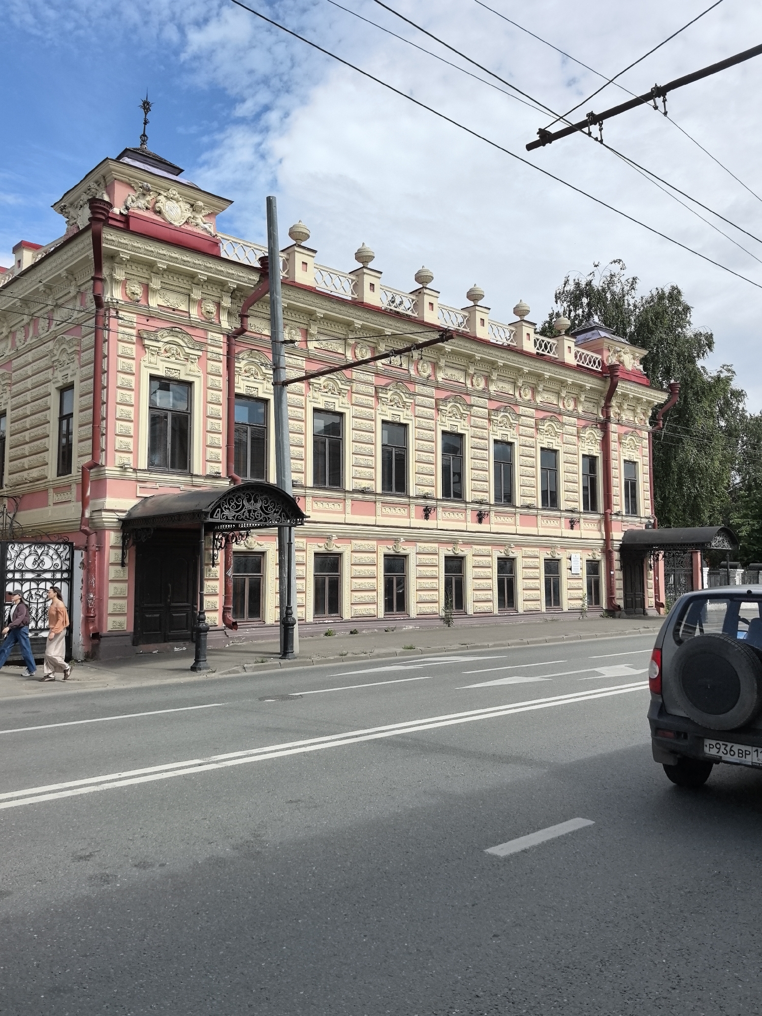This media file captures a street view in Usady, Russia, under a partly cloudy sky. The scene features an ornate, multi-story building with a pink and cream facade, characterized by detailed architectural embellishments, including decorative window frames, cornices, and a small turret with a spire. The building appears to be a historic structure, possibly a mansion or government building, with multiple windows and several entrances.
In the foreground, a paved road with white lane markings runs horizontally across the frame. A dark-colored SUV with a spare tire mounted on the back is visible on the right side of the road, partially obscuring the building's right side. On the sidewalk in front of the building, two individuals are walking from left to right. One is wearing a light-colored outfit, and the other is in darker clothing. Overhead, electrical wires for what appear to be a tram or trolley system crisscross the sky, suggesting a public transportation route.
Notable details include the architectural style of the building, the presence of overhead wires, and the general appearance of a town or city street. The lighting suggests it is daytime, with a mix of sun and clouds. No specific events are depicted, but the scene portrays a typical day on a street with historical architecture and active traffic. There is visible text on the license plate of the SUV, which reads "P936 BP 11".