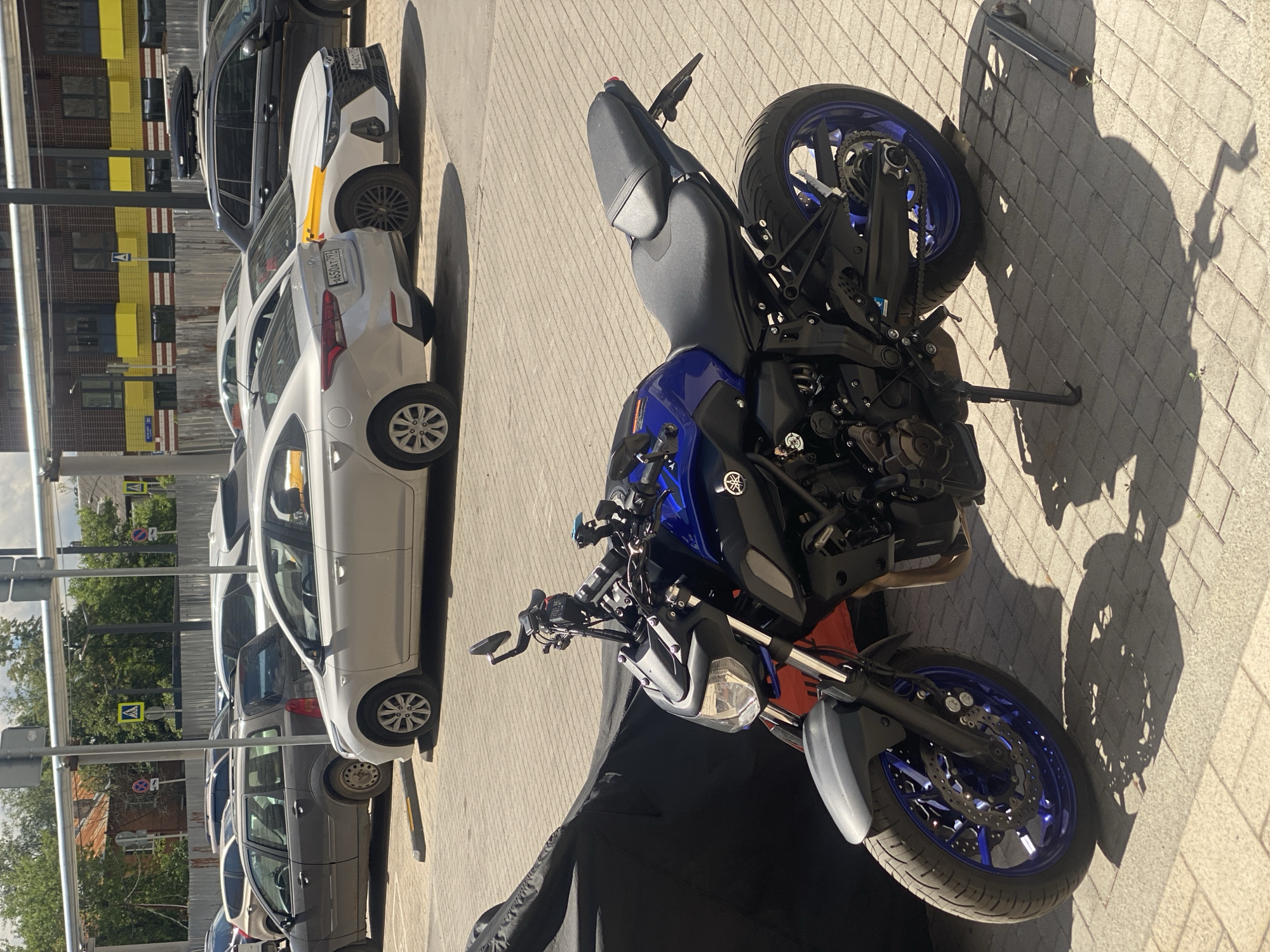 A blue and black motorcycle is parked on a paved area next to several cars. The motorcycle is in the foreground, with its front wheel facing right. It has a sporty design with exposed engine parts and a blue rim on the rear wheel. A black motorcycle cover is partially visible in front of the motorcycle.
To the left of the motorcycle are several cars parked in a row. The closest car is white, with its rear end facing left. Further down the line are a dark gray car and other vehicles. The cars are parked on a paved surface that appears to be a street or parking lot.
In the background, there is a building with yellow and brown colored panels. Metal pipes run along the side of the building. Trees and some signage are also visible.
The scene is illuminated by sunlight, casting shadows on the pavement. The time of day appears to be daytime, likely morning or afternoon, given the strong shadows. The weather seems pleasant, with clear skies implied by the bright sunlight. The location context is Kotelniki, Russia. No people are visible in the image. There is some text visible on the license plates of the cars, but it is not legible. A logo, possibly of a manufacturer, is visible on the side of the motorcycle.