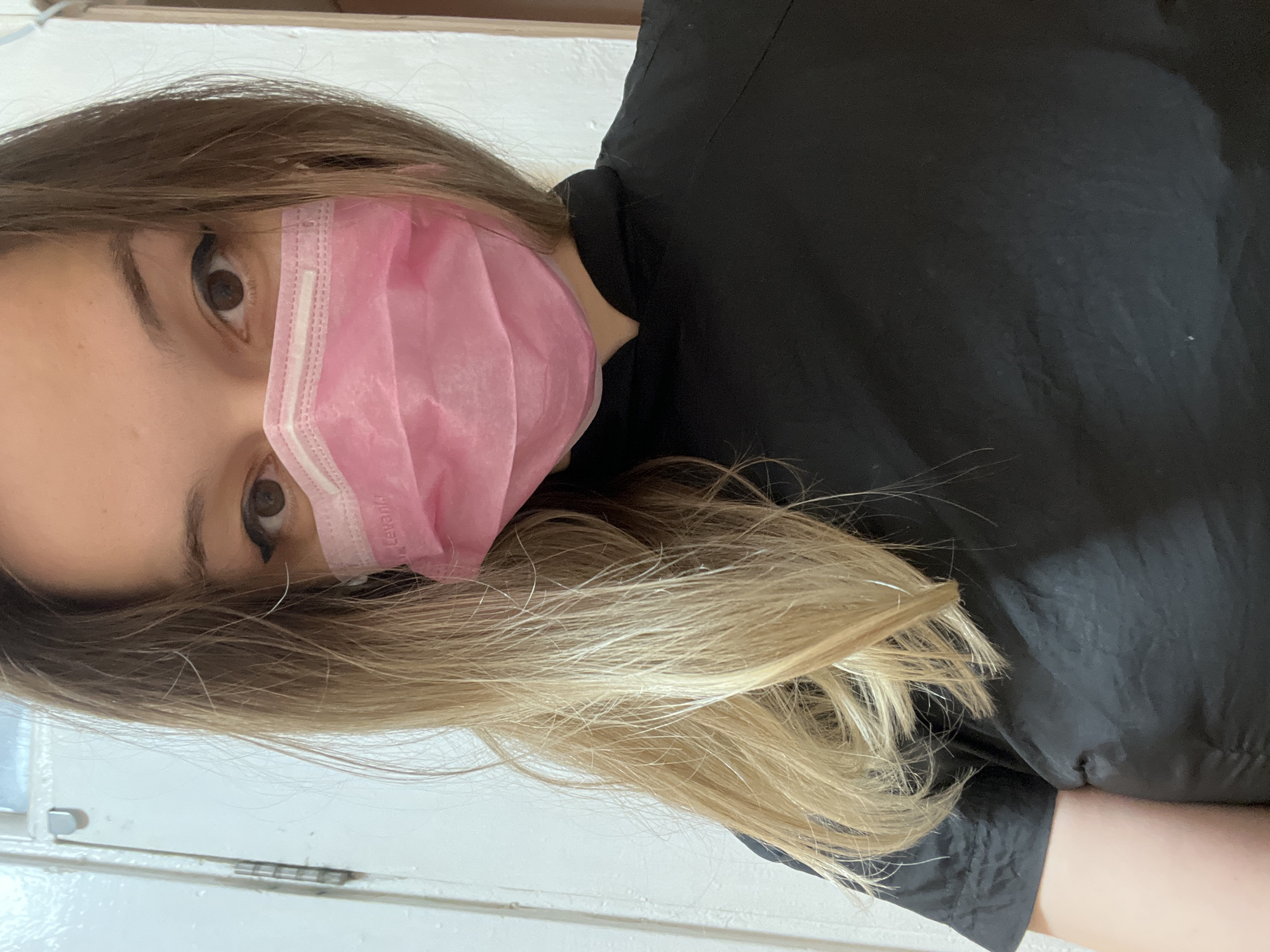 A medium close-up shot shows a young woman with blonde and brown hair, wearing a pink face mask and a black shirt. She is looking directly at the camera with brown eyes. Her hair is long and loose, falling over her shoulders. The background is a white wall with a door on the left side. The lighting is natural, coming from the front.