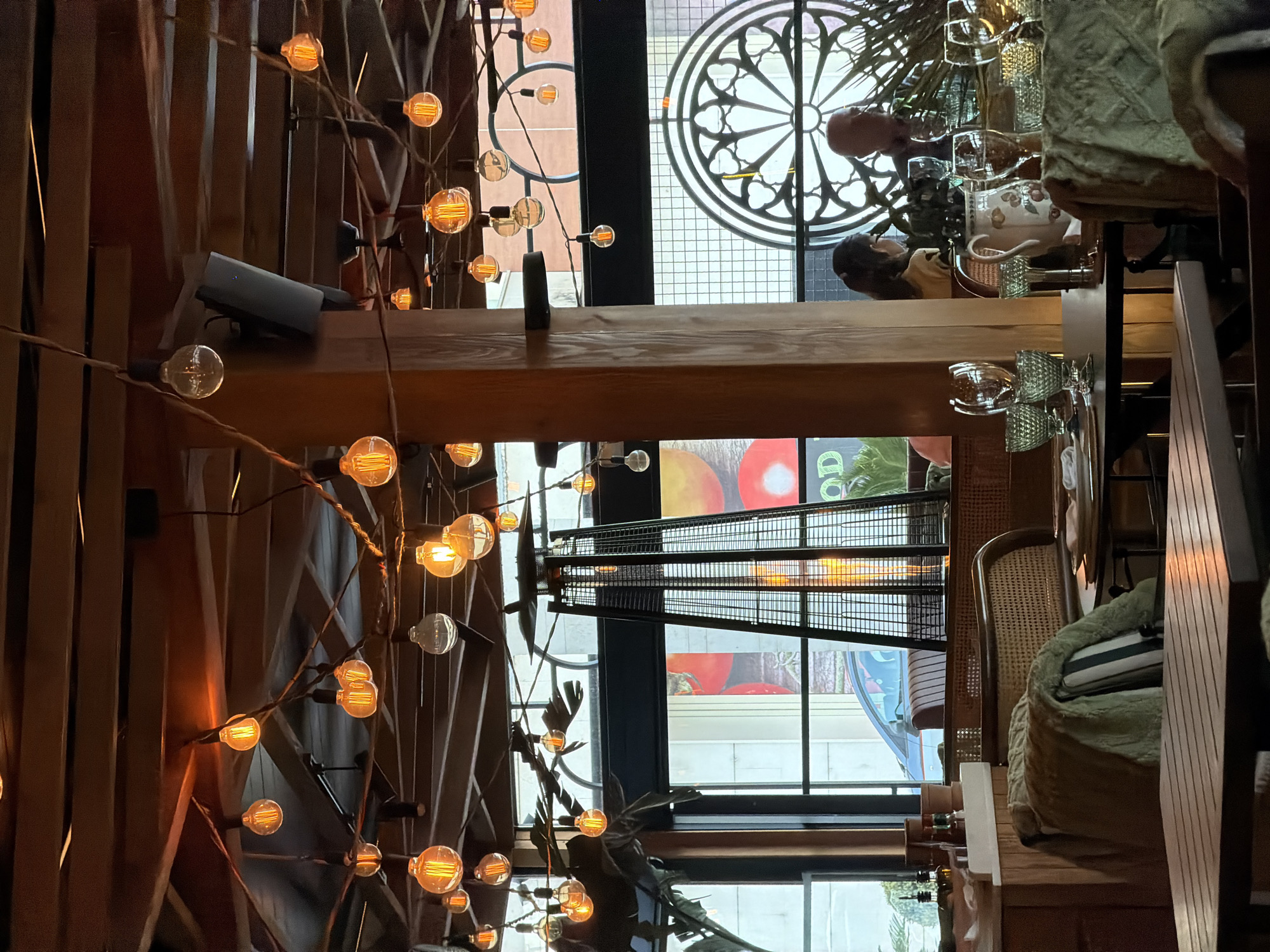 The image depicts the interior of a restaurant or café in Budva, Montenegro. The space has a rustic, urban design aesthetic, with exposed wooden beams and a decorative lighting fixture featuring numerous Edison-style bulbs strung across intertwined branches. A mezzanine level is visible, and on it are seated patrons at tables. A spiral staircase leads to the upper level. The exterior features windows with decorative metal grilles or patterns. From the windows, parts of buildings are visible, suggesting the location is in a fairly urban or commercial area of the city. The lighting is warm, creating a cozy atmosphere. Details like chairs with cushions and dining setups suggest a focus on customer comfort.