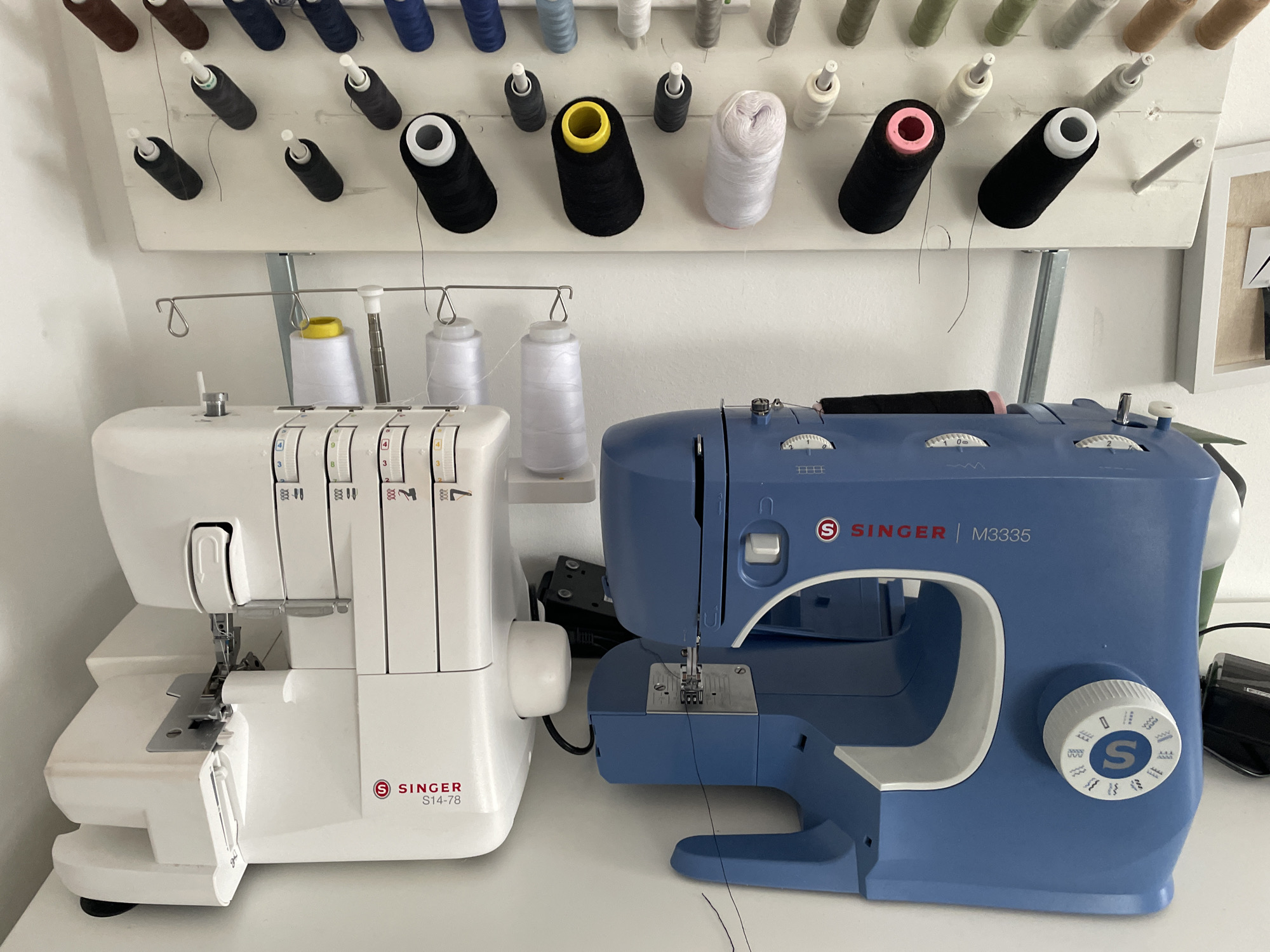 The image depicts a sewing room, likely in a residential setting, featuring two sewing machines and numerous spools of thread organized on a wall-mounted rack. The sewing machines are positioned on a white surface, possibly a table or counter. To the left is a white serger sewing machine, identified by the "SINGER S14-78" logo. To its right is a blue Singer M3335 sewing machine. Both machines are equipped with spools of thread.
Above the machines, a white wooden rack holds an array of spools of thread in various colors, including black, white, pink, brown, blue, and green. The threads are arranged in neat rows. Some spools have distinct colored caps, such as yellow and pink.
The environment appears to be an indoor space, likely a dedicated crafting area. The lighting suggests an ambient indoor setting, possibly with natural light contributing. There are no individuals present in the image. The overall impression is one of an organized and functional sewing space. The specific location in Nidda, Germany, is not discernible from the visual information presented in the image itself; this context would require external knowledge or metadata.