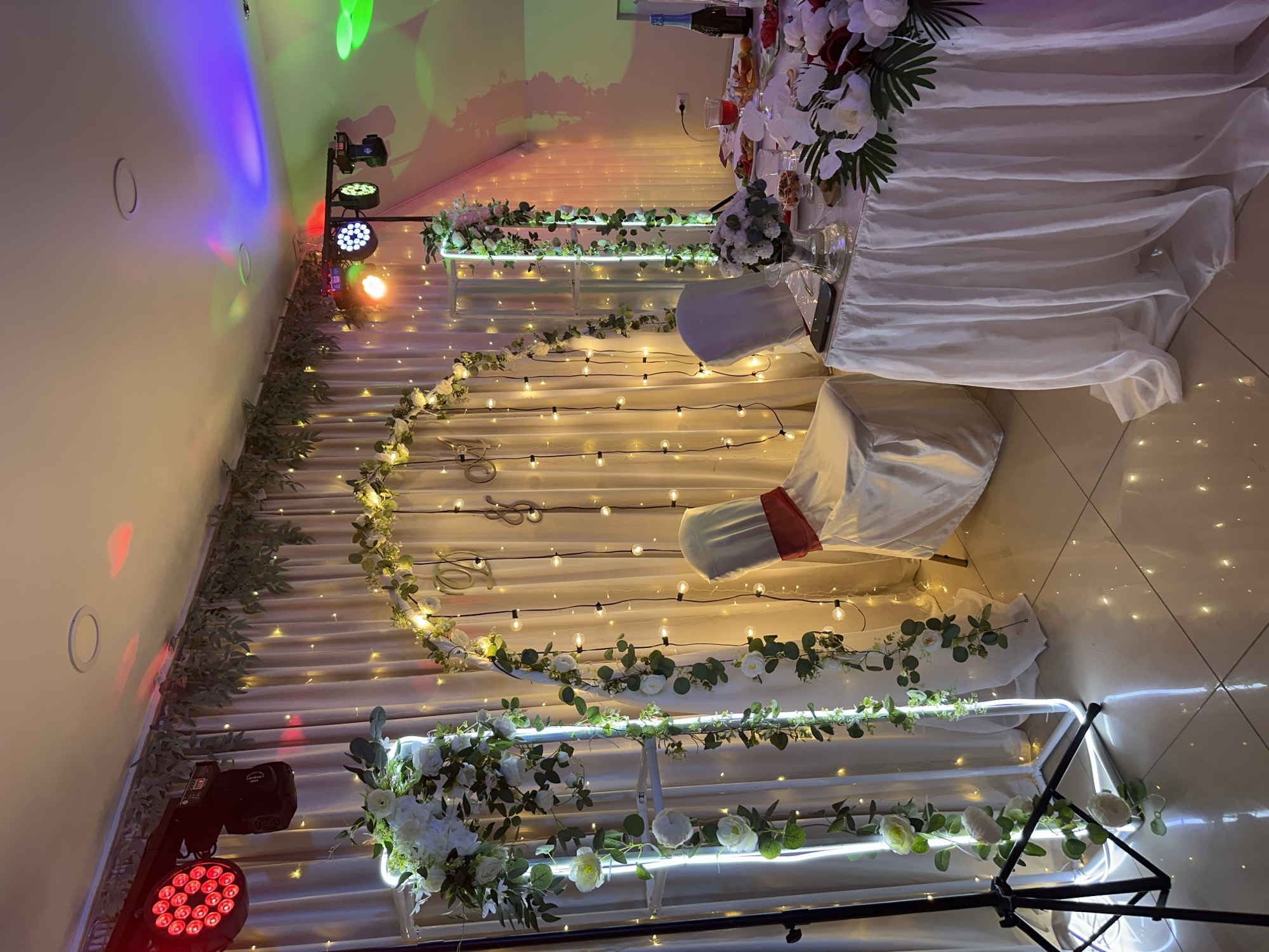 This media file captures a decorated wedding reception setup, likely in Chaikovsky, Russia. The scene is dominated by draped white fabric, creating a backdrop for the event. A long table, draped in white, is adorned with floral arrangements featuring white and red flowers and lush green foliage. In the foreground, string lights with warm yellow bulbs are draped across the fabric, creating a festive and intimate ambiance.
Further back, a multi-tiered structure, also decorated with greenery and white flowers, is illuminated by integrated LED lighting. Two chairs are visible, one draped in white fabric with a red sash. Several spotlights, including some with red and colored lights, are positioned around the room, suggesting that the event is either ongoing or about to begin. The lighting creates colorful reflections on the wall and ceiling.
The overall atmosphere appears to be celebratory and elegant, with the decorations emphasizing a romantic theme. The time of day is not explicitly shown, but the indoor setting and artificial lighting suggest it could be evening or a dimly lit indoor venue. There is no visible text in the media file.