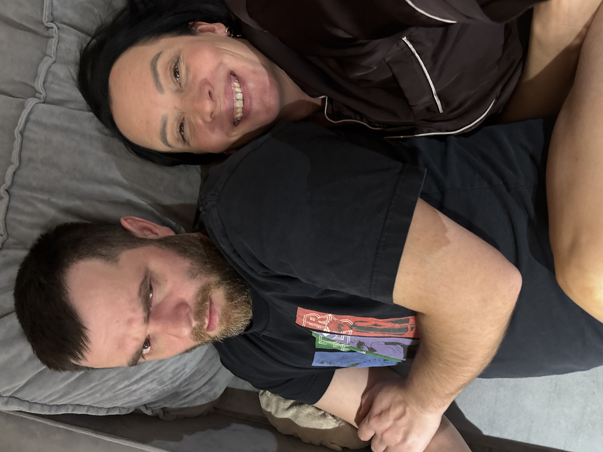 A woman is lying on top of a man on a gray surface, possibly a bed or couch. The woman has dark hair, fair skin, and is smiling broadly. She is wearing a dark-colored, satin-like top with white trim. The man has a beard, fair skin, and is wearing a black t-shirt with a colorful graphic design. His expression is more serious. The background consists of the gray surface they are lying on. The location is not specified.