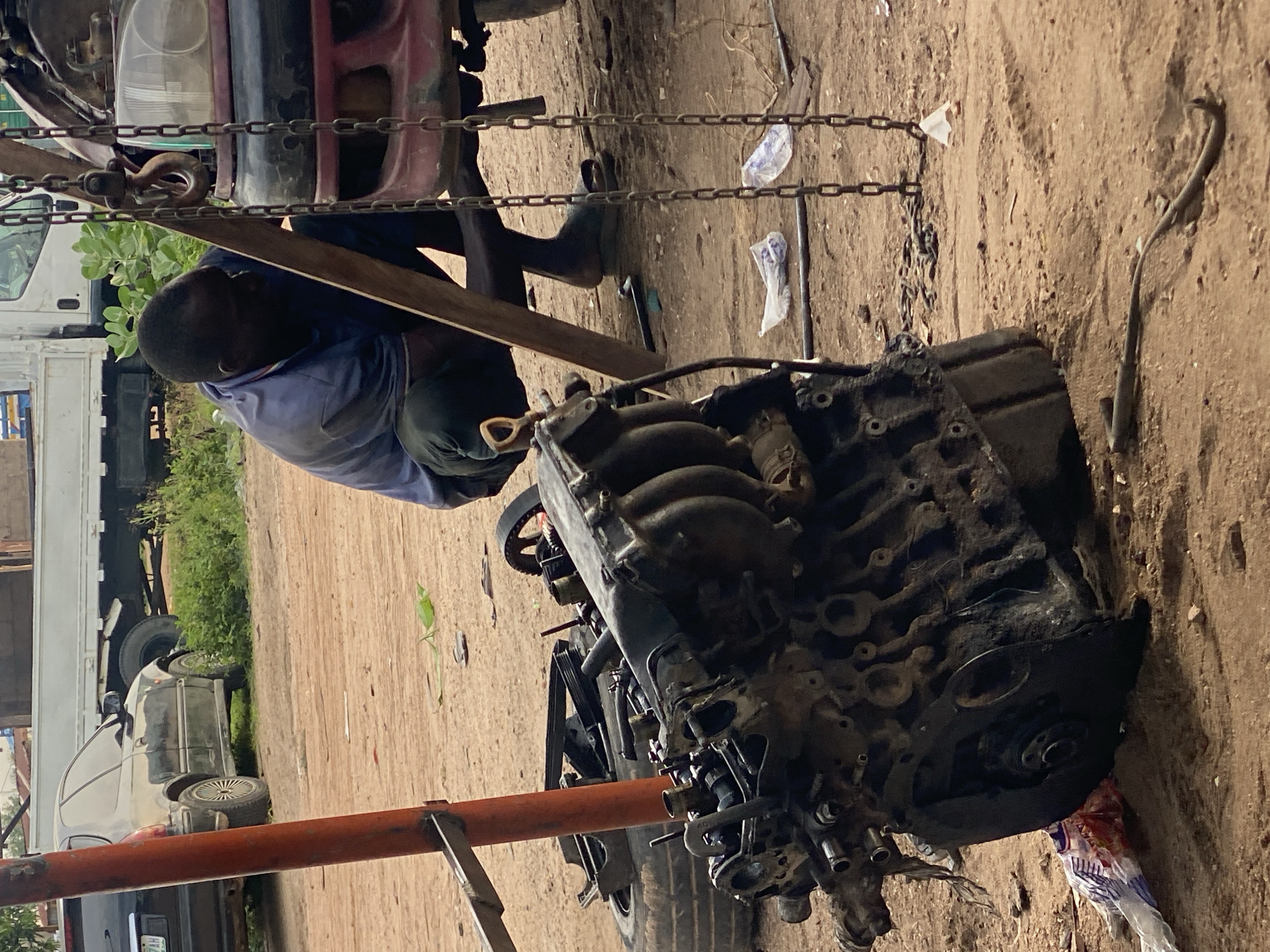 The image depicts an outdoor auto repair scene, likely in Jalingo, Nigeria. A dark-skinned man, dressed in a light blue shirt and dark pants, is actively engaged in working on a vehicle. He appears to be crouched or kneeling, with his body angled towards the engine of a red vehicle suspended by chains and a wooden beam. A large, dirty engine block rests on the ground in the foreground, suggesting it has been removed from a vehicle. The ground is a sandy, dirt surface, indicative of a workshop or open-air repair area. In the background, a black car and the front of a white truck are partially visible. The overall environment suggests a busy, functional, and likely modest repair shop.