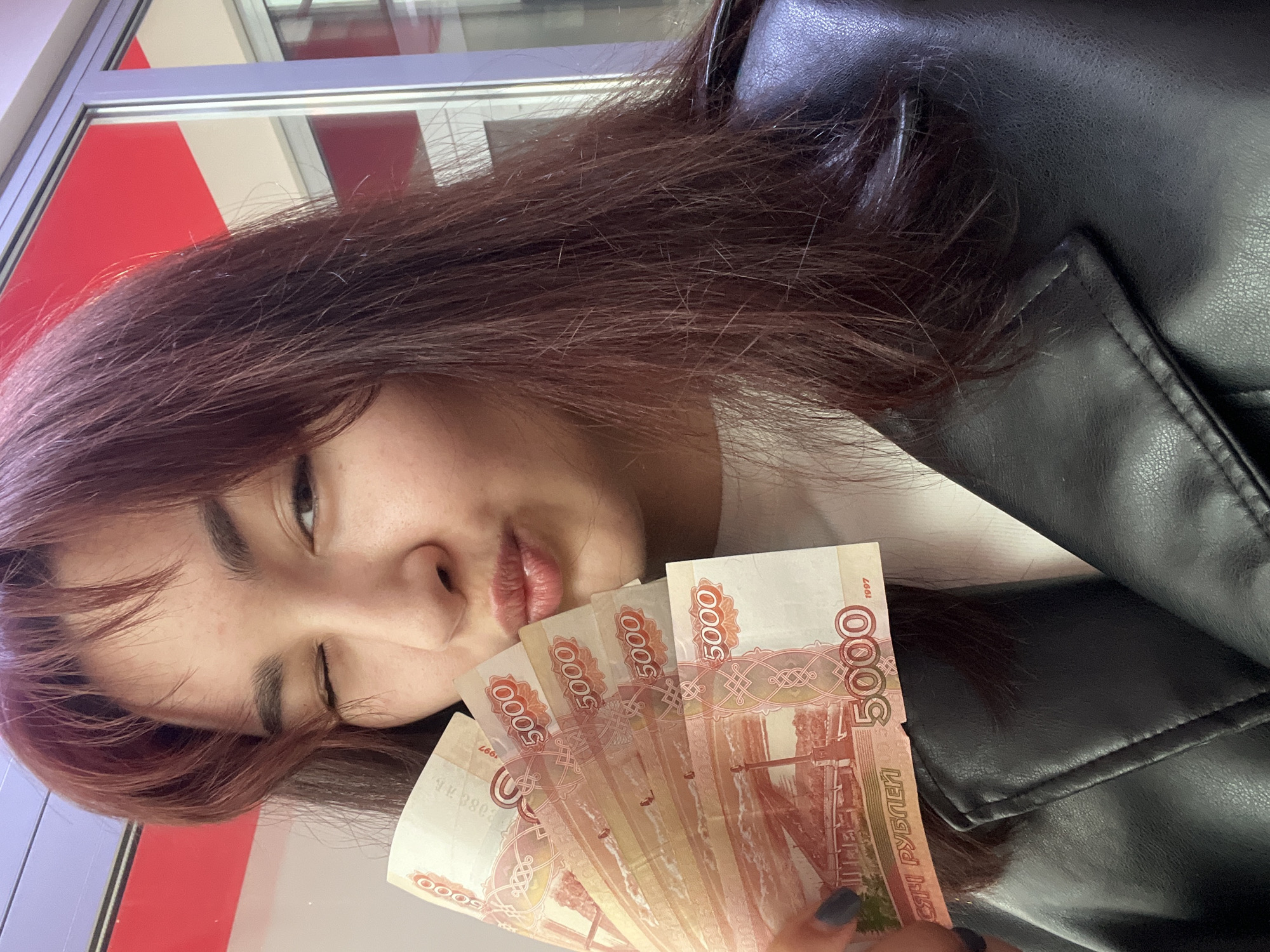 A young woman poses for a selfie, holding several 5000 Russian Ruble banknotes near her face. She is partially winking and pursing her lips. She is wearing a black leather jacket and a white shirt, and her hair is dark brown with reddish highlights. The background shows a window with red and white panels, suggesting an indoor location with a view of the outside. While it isn't directly apparent from the image, it is specified in the prompt that the location is in Neftekamsk, Russia.