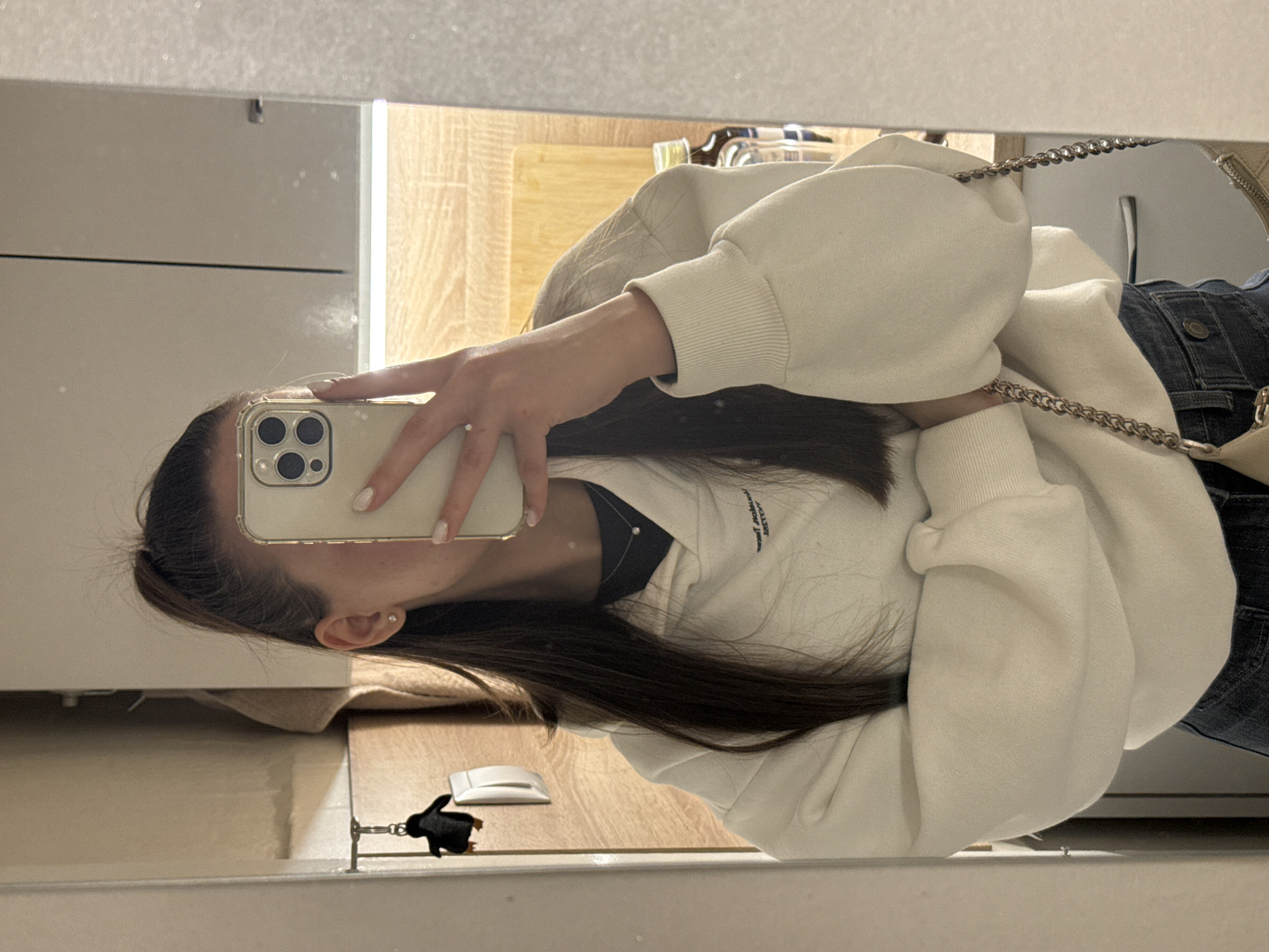 A young woman is taking a mirror selfie with an iPhone 13 or 14 series phone, evident by the 3-lens camera design. She is wearing a white sweatshirt and jeans. A small purse with a chain strap is visible. The reflection shows a bathroom or similar room, with white cabinets, wooden floors, and a small decorative object resembling a penguin. A phone is on the counter, next to the penguin, indicating they may be hotel phones. The image does not provide sufficient information to determine a specific location within Sofia, Bulgaria.