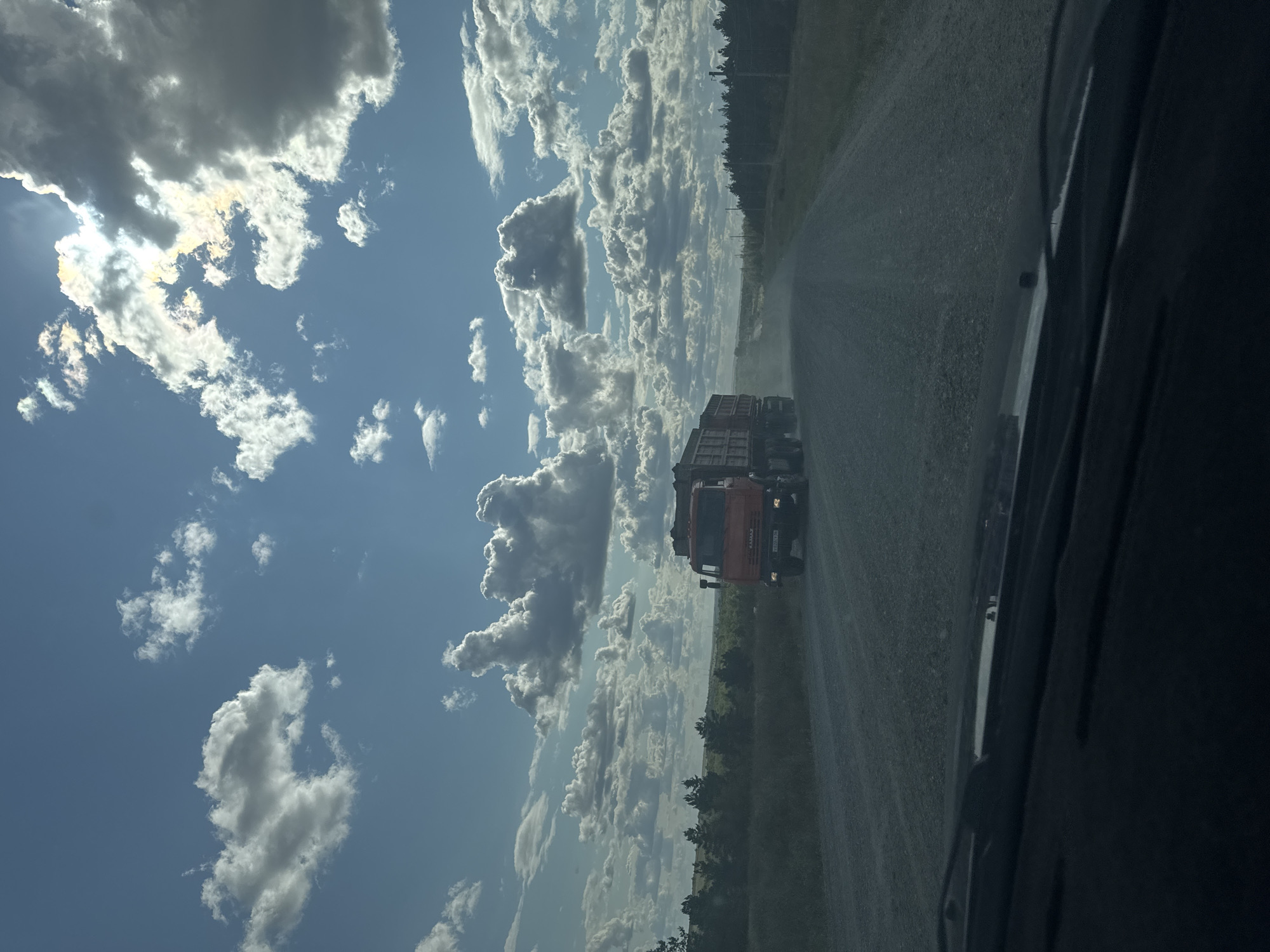 In Vasilievka, Russia, a bright, cloudy sky looms over a dirt road. A red KAMAZ dump truck, loaded with dark material, travels away from the camera, kicking up a trail of dust. The truck's headlights are on. The road is flanked by sparse trees and grassy fields on either side. The scene is captured from inside a vehicle, with the dashboard and windshield visible in the foreground.