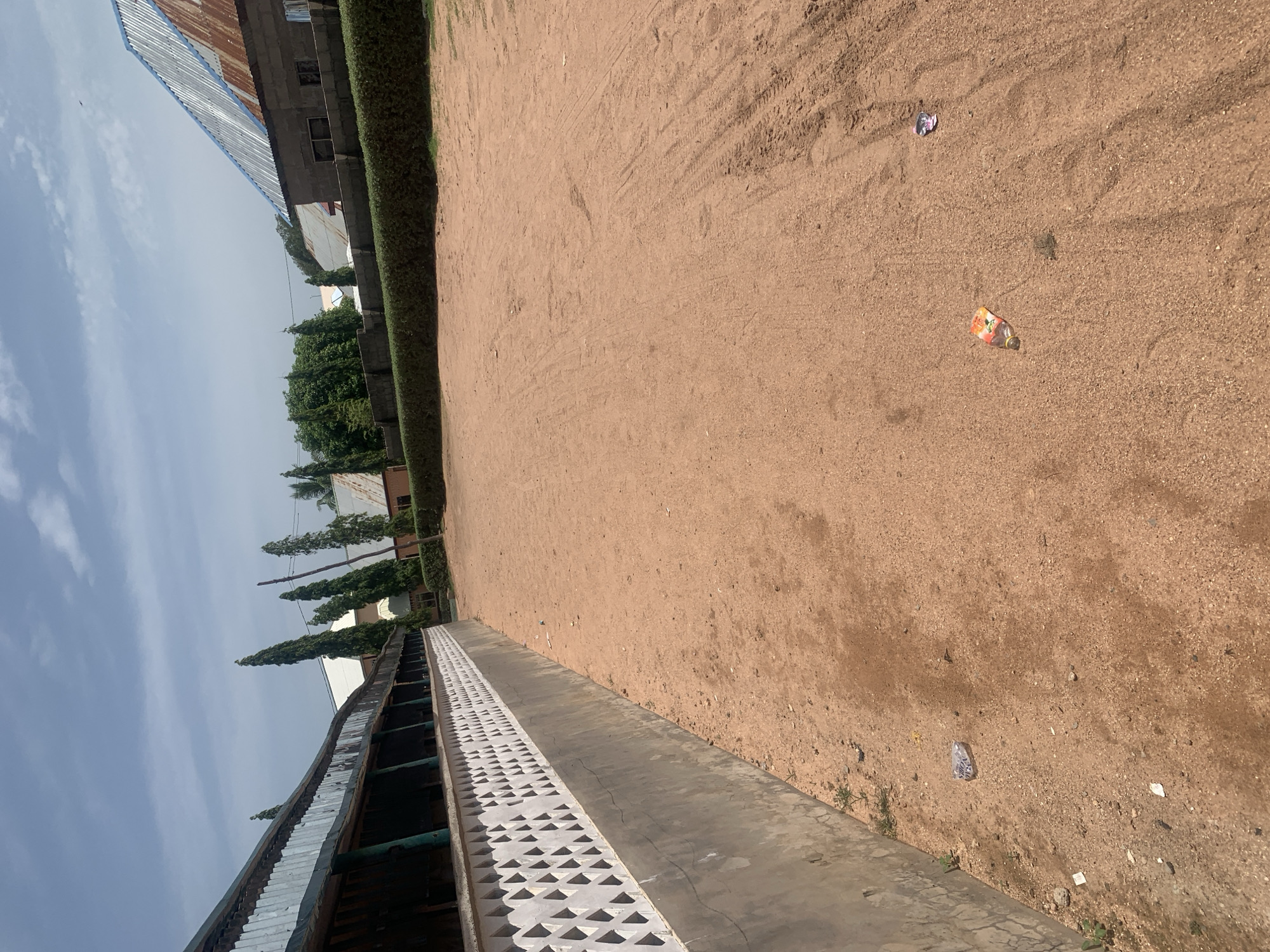 The image depicts an outdoor scene in Jauro Yinu, Nigeria. A long, weathered structure with a corrugated metal roof runs along the left side of the frame. Below the roof is a white decorative wall with diamond-shaped openings, followed by a gray concrete base. The ground is a light brown, sandy dirt surface, with some tire tracks visible in the distance. In the foreground, on the sandy ground, there are a few pieces of litter, including a plastic bottle with a colorful label and a crumpled plastic wrapper. In the background, beyond the structure, are buildings with corrugated roofs and some trees. The sky is blue with scattered white clouds.
