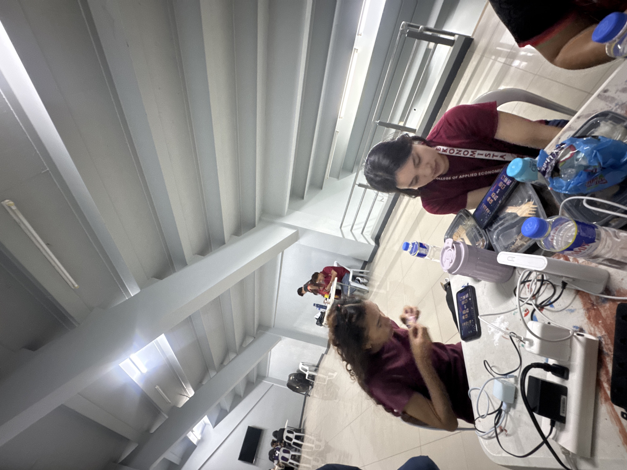 The image shows an interior view, potentially an atrium or large waiting area, likely in Davao City, Philippines. The space is brightly lit with a high, ribbed ceiling and white supporting columns. In the foreground, two women are seated at tables. One woman with curly hair is looking at a mobile phone, while the other woman, wearing a maroon top with the word "ECONOMISTA" printed on the straps, is looking at a laptop or tablet. The tables are cluttered with water bottles, food containers, cables, and electronics. In the background, additional people are seated at tables in the distance. The overall impression is one of a busy, informal study or work area.