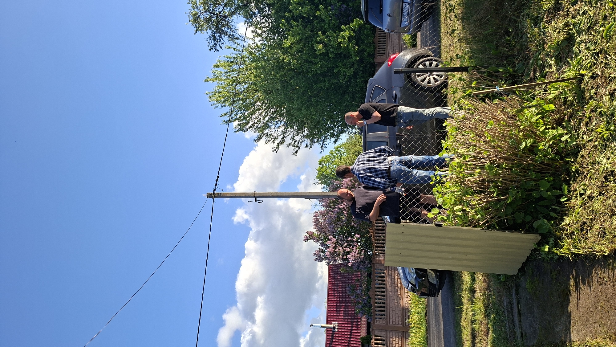 The image depicts an outdoor scene in Сваткаўскі сельскі Савет, Belarus, under a bright blue sky with scattered white clouds. Three men are standing near a chain-link fence, seemingly engaged in conversation or observation. One man is wearing a black t-shirt, another a blue and white plaid shirt, and the third also wears a black t-shirt. Behind them, a dark-colored vehicle is partially visible. To the right of the men, on the other side of the fence, is a bush with green and brown foliage. In the background, a red-tiled roof and a brick wall can be seen, along with a lilac bush and trees. An electric pole with wires extending across the sky is also visible in the foreground. A sheet of corrugated metal is leaning against the fence. Grass and other foliage line the ground.