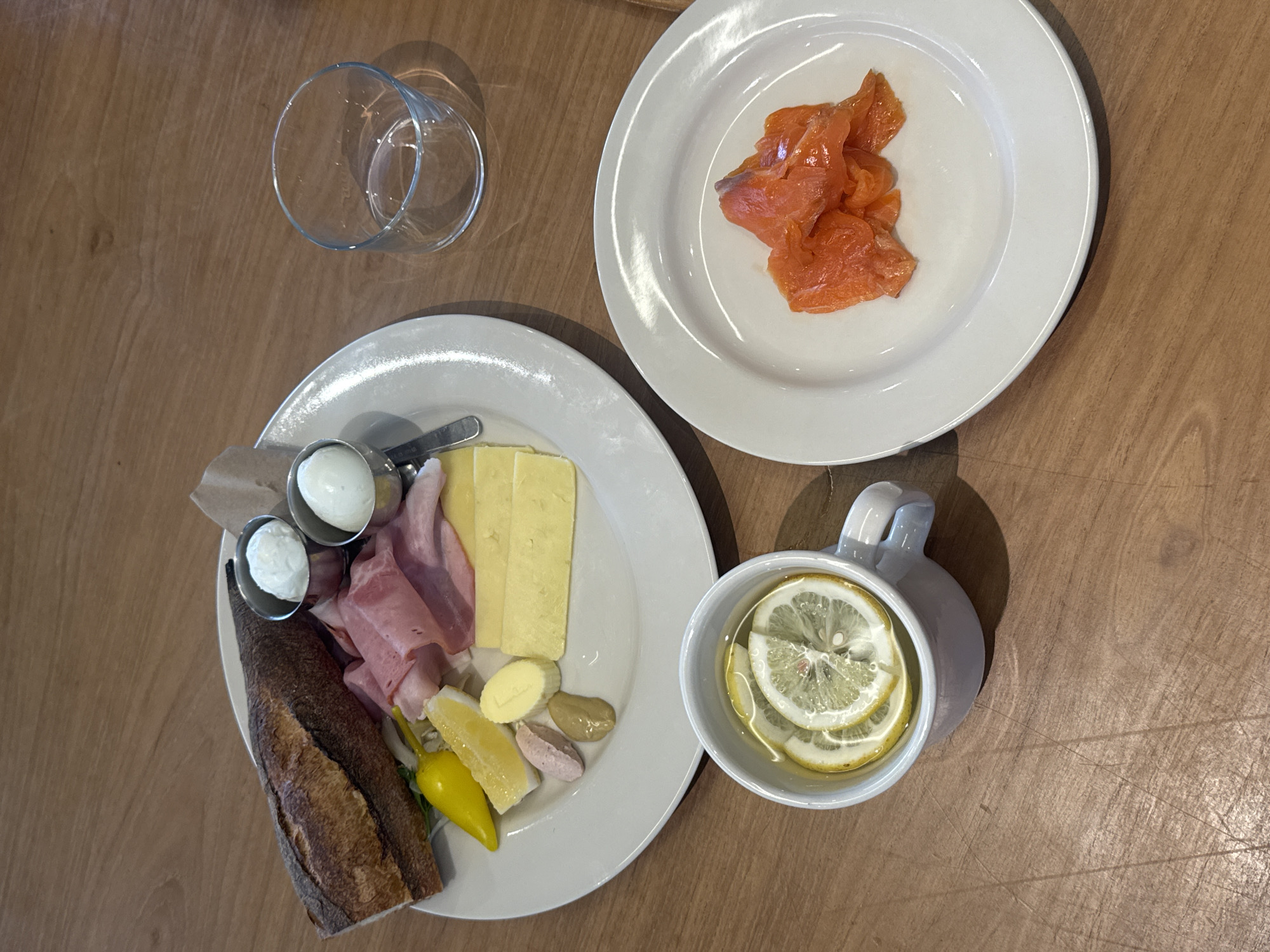 A top-down shot shows a table setting with various food items. On the table are two white plates, one with smoked salmon and another with a baguette, ham, cheese, eggs, and peppers. There is also a glass and a mug filled with a liquid containing lemon slices. The location is identified as North Kuta, Indonesia.