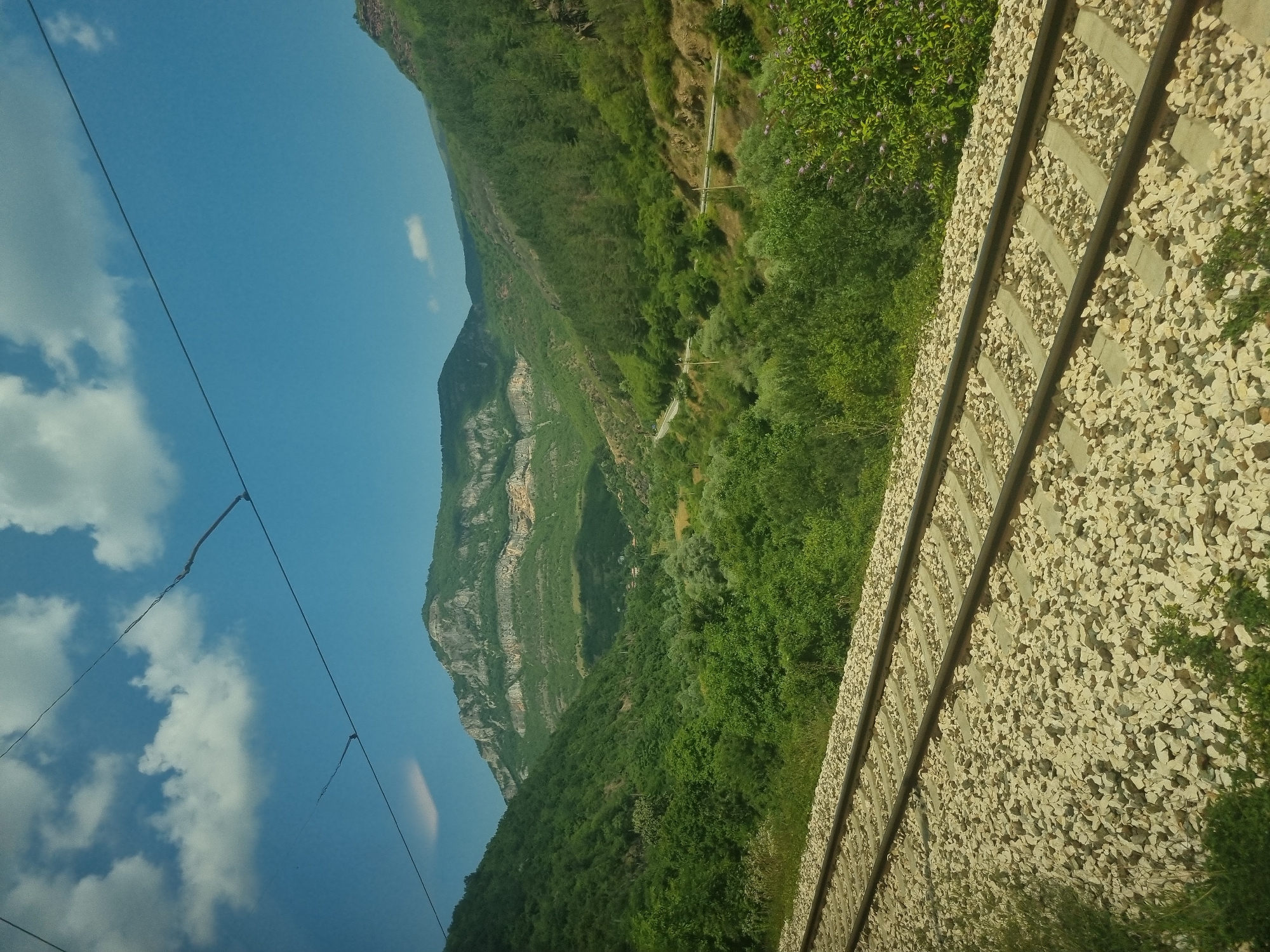 The image depicts a scenic view of a mountain valley with a train track in the foreground. The train track, composed of dark rails and light-colored ballast, runs diagonally across the lower portion of the frame. Beyond the tracks, the landscape is dominated by lush green trees and vegetation, indicating a rural or natural setting. In the mid-ground, a road winds its way through the valley, bordered by trees on either side. The background features a majestic mountain range with layered rock formations and vegetation. The sky above is a clear blue, dotted with scattered white clouds. The overall impression is one of natural beauty and tranquility. The provided location of Ochindol, Bulgaria, suggests this is a view from or near that area. There are no people or animals visible in the image. The scene is captured in daylight, with clear visibility and natural lighting.