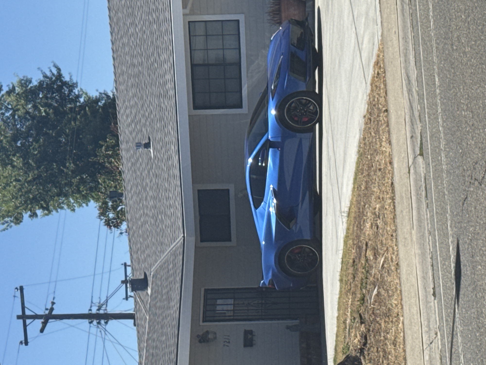 A blue sports car is parked in the driveway of a single-story house. The house is light-colored and has a gray shingled roof. There are two windows visible on the front of the house, one to the left of the car and one to the right. A security door is in front of the entrance to the house. A telephone pole with power lines is visible to the left of the house, and a large tree is to the right. The car appears to be a Chevrolet Corvette. The scene is outdoors on a sunny day, with a clear blue sky. The car is parked on a paved driveway that extends to the street. A patch of dry grass and leaves is located between the driveway and the street. The house number "7211" is visible on the wall near the entrance.