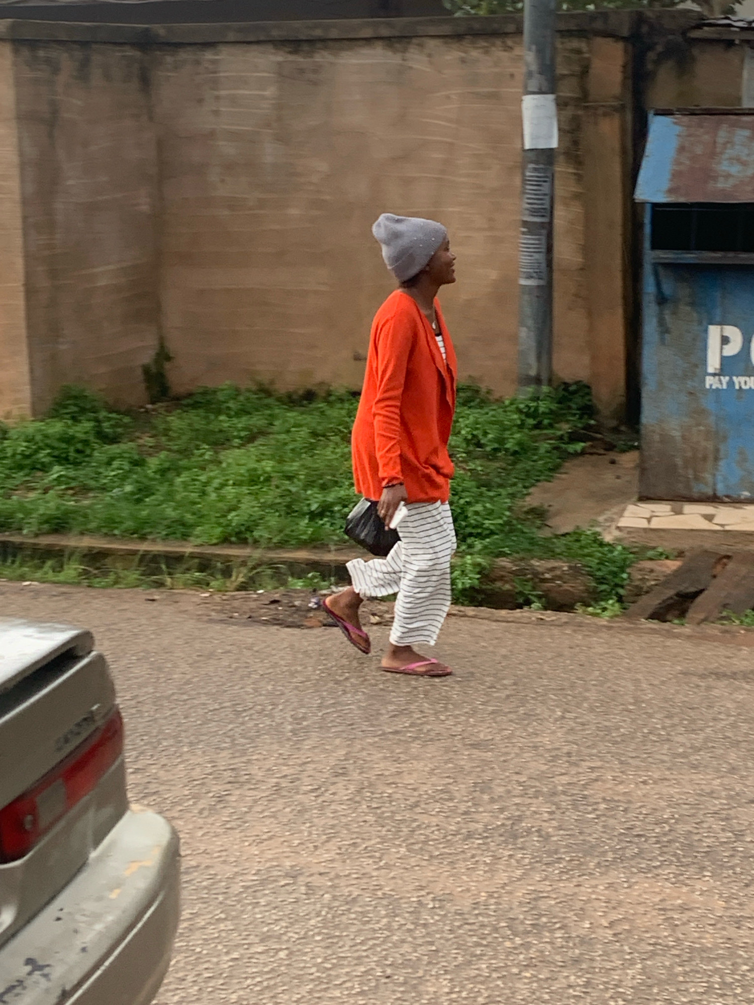 A medium shot shows a person walking on a paved road in Jos, Nigeria. The person is wearing a grey beanie, an orange cardigan, and striped pants, with pink flip-flops on their feet. A black purse is slung over their shoulder. To the right of the person is a blue metal structure with "PAY YOUR" visible in white lettering. In the background, a tan-colored wall with a dark coping line runs horizontally. Green vegetation is visible between the wall and the road. To the left of the frame, the rear of a silver car is partially visible, with its red taillight illuminated. The sky is overcast.