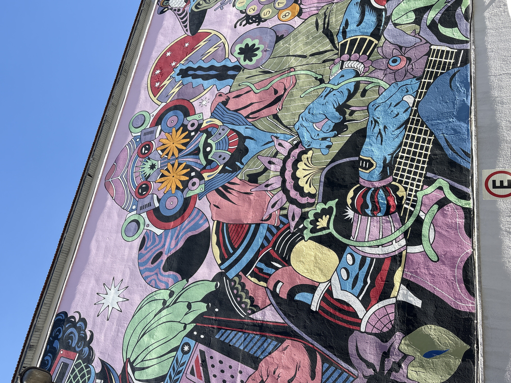 This is a photograph of a large, colorful mural painted on the side of a building in São Paulo, Brazil. The mural features a stylized, abstract figure with multiple eyes and decorative elements. The figure appears to be holding a guitar or similar stringed instrument. The artwork is characterized by bold lines, vibrant colors such as pink, blue, green, yellow, and orange, and a complex, psychedelic design. The mural covers a significant portion of the building's facade, which is painted a light pink. A clear blue sky is visible above the building, and the edge of the roof and gutters are also visible. At the bottom of the image, a white wall with some texture and a red and white circular sign with the letter "E" are visible. The mural is likely located in an urban area of São Paulo, given the presence of the building and the street signage.