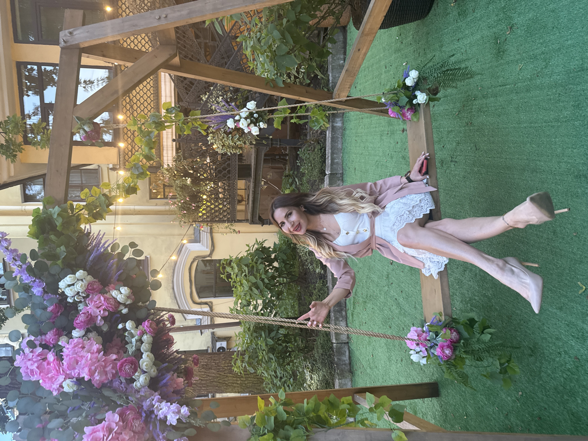 An outdoor scene in Saint Petersburg, Russia, features a woman seated on a wooden swing, decorated with abundant artificial floral arrangements in shades of pink, white, and purple, interspersed with green foliage.
The woman, with long ombré hair and full makeup, is smiling towards the camera. She wears a white sleeveless top with lace detailing, a light pink blazer, and nude high-heeled pumps, with her legs crossed at the ankles. Her left hand grips a swing rope, while her right hand holds a red smartphone resting on the wooden seat.
The ground beneath the swing is artificial green turf. The background consists of a yellow stucco building featuring arched windows, ornate architectural details, and string lights strung across the upper facade. Dense green bushes and a dark wrought iron balustrade are visible behind the swing structure. A tree trunk and an exterior air conditioning unit are also discernible on the far left.