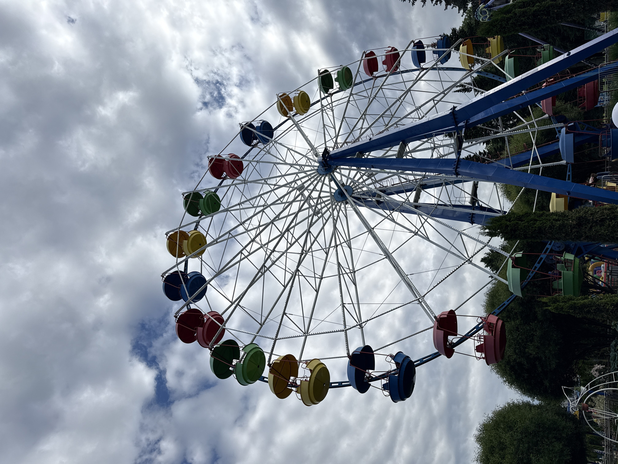 An outdoor, eye-level, low-angle shot captures a large, colorful Ferris wheel against a cloudy sky. The Ferris wheel is predominantly white and blue, with a variety of brightly colored gondolas, including red, yellow, green, and blue, suspended from its structure. The gondolas appear to be empty. The Ferris wheel is supported by substantial blue metal legs.
The surrounding environment includes lush green trees and foliage, suggesting a park or amusement area. In the lower left corner of the image, part of another white structure, possibly a smaller ride, is visible. The ground area appears to be paved or gravel.
The image was taken on a day with a heavy cloud cover, with patches of blue sky peeking through. The lighting is diffused, consistent with an overcast sky. There are no people visible in the frame, and no obvious actions are taking place. The scene conveys a sense of stillness, perhaps indicating the park is not yet open or is in a quieter period.
This scene is located in Saint Petersburg, Russia, at the "Divo Ostrov" amusement park, situated on Krestovsky Island.