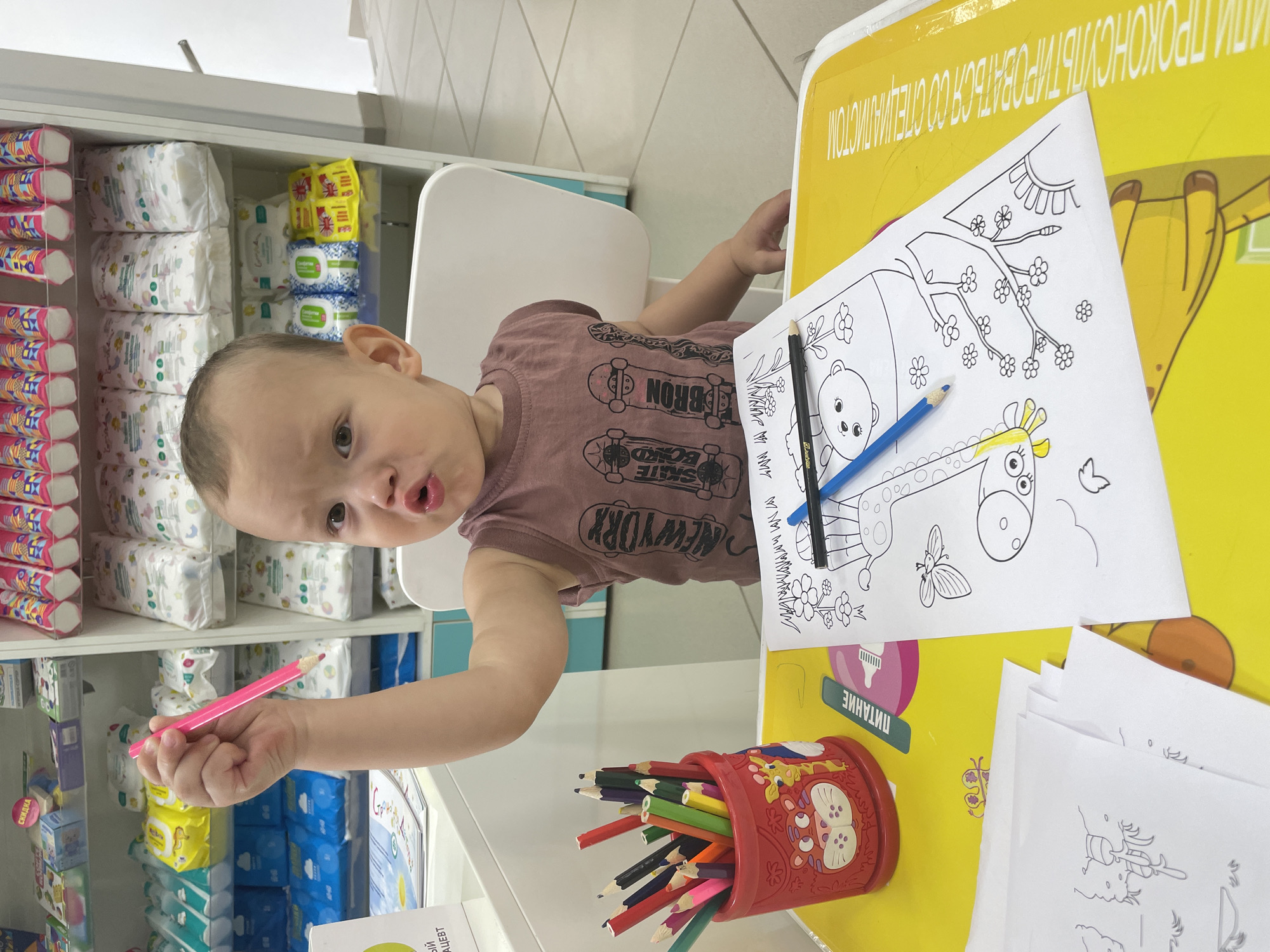 In a retail store, possibly a pharmacy or baby supply store, a young child sits at a yellow table with coloring pages and a red pencil holder filled with colored pencils. The child is holding a pink pencil and appears to be looking at the camera with a surprised expression. Behind the child, shelves are stocked with diapers and other baby products. The store appears to be clean and well-organized. The presence of Russian text on some of the products and signage suggests the location is in Russia.