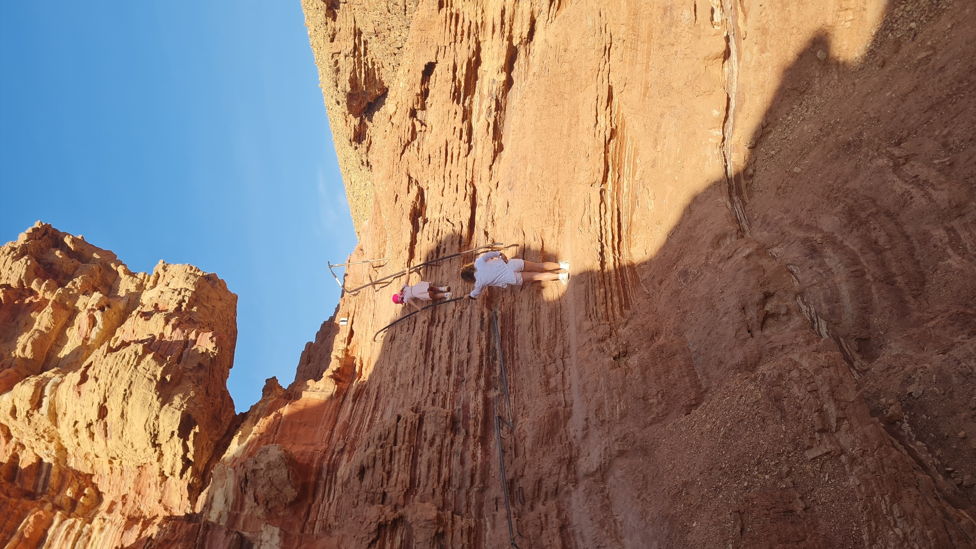 The image shows a low-angle shot of a reddish-brown rock formation in Hevel Eilot Regional Council, Israel. Two people are standing on a metal walkway or steps built into the cliff face. One person, further along the walkway, is wearing white clothing. The other person, closer to the foreground, is wearing a pink hat and shorter, light-colored clothing. The walkway ascends the rock face. The sky is visible above the rock formation, and a large shadow is cast onto the rock to the right of the walkway.