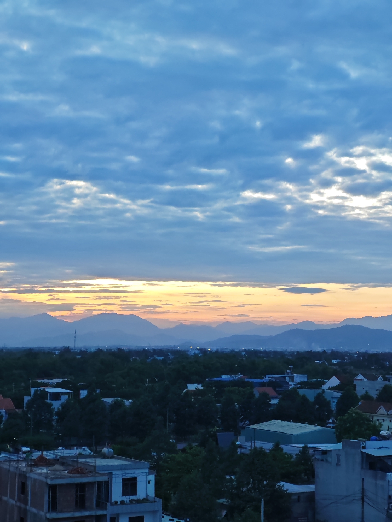 The image depicts a cityscape at dusk or dawn, overlooking a mountain range in the distance. The sky is filled with layered clouds, with a band of orange and yellow light near the horizon, indicating either sunrise or sunset. Below the sky, a dense canopy of trees covers the midground, interspersed with buildings of varying sizes and styles, characteristic of a developing urban area. In the foreground, construction is visible on a multi-story building, with exposed concrete and brickwork, suggesting ongoing development. The overall scene suggests a view from a slightly elevated position, looking out over a suburban or semi-urban area of Da Nang, Vietnam.
