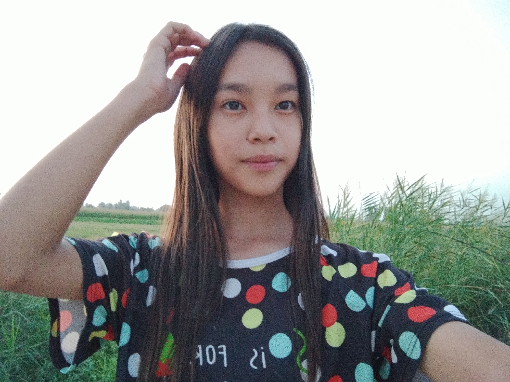 A young girl with long, dark hair is taking a selfie outdoors. She is wearing a dark t-shirt with colorful polka dots. Her left arm is raised, and she is touching her hair. The background consists of a field and tall grass, suggesting a rural or natural setting. The sky is bright, indicating it is daytime, possibly late afternoon given the warm lighting. The overall mood appears relaxed and casual. The location is identified as Kochkor-Ata, Kyrgyzstan. There is some visible text on the girl's shirt, but it is partially obscured and difficult to read clearly.