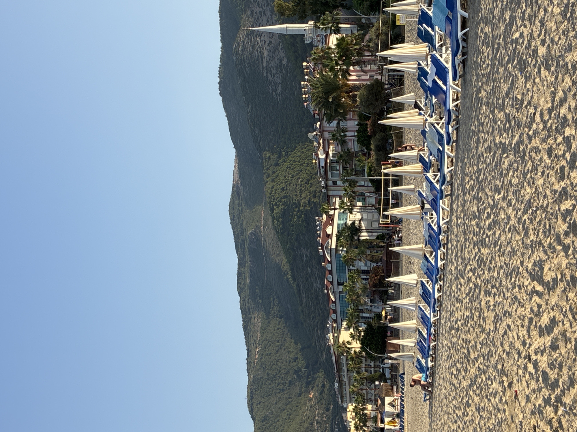 This is a photograph of a beach in Alanya, Turkey, with the city's characteristic coastal mountain range in the background. The foreground is dominated by a wide expanse of sandy beach, with numerous rows of blue and white lounge chairs and white beach umbrellas set up. In the mid-ground, there is a line of buildings that appear to be hotels and resorts, with palm trees interspersed throughout. A white minaret, characteristic of a mosque, is visible rising above the buildings. One person is seated on a lounge chair on the left side of the frame. The sky is clear and blue.